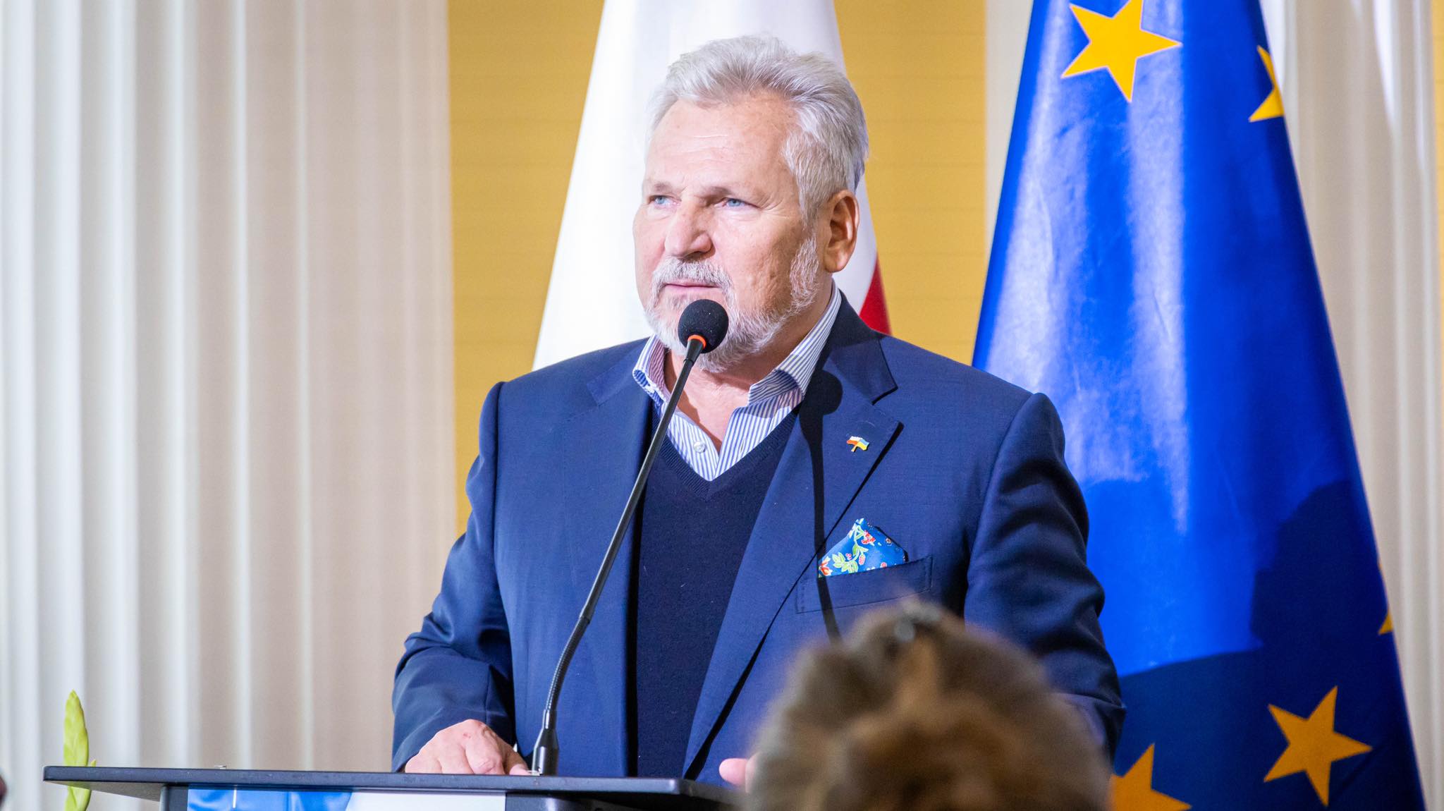 Na zdjęciu widać mężczyznę w garniturze, który przemawia z mikrofonem, w tle znajdują się flagi Polski i Unii Europejskiej.