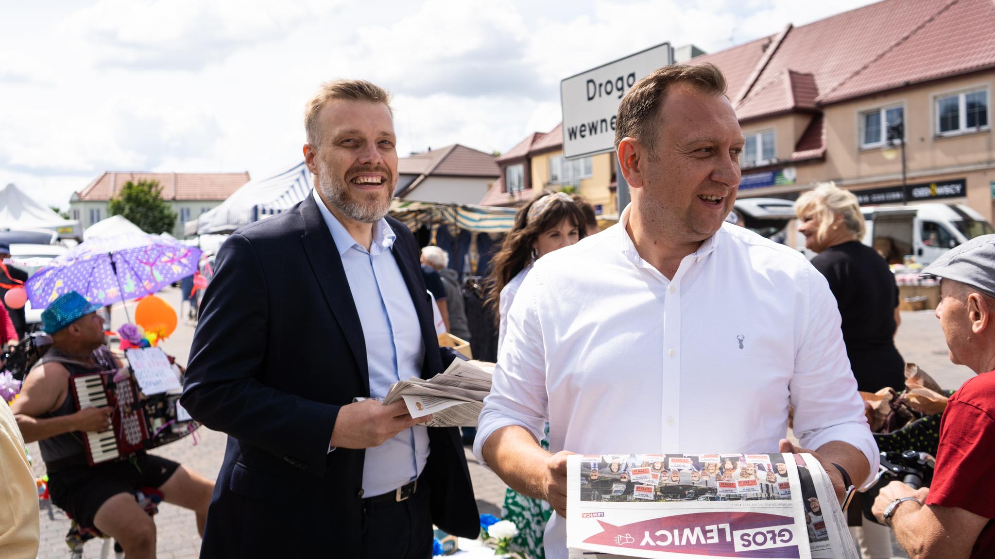 Na zdjęciu widać dwóch mężczyzn śmiejących się i trzymających gazety, przechodzących obok stoiska na lokalnym rynku, w tle widać kolorowe parasole oraz kilku innych ludzi.