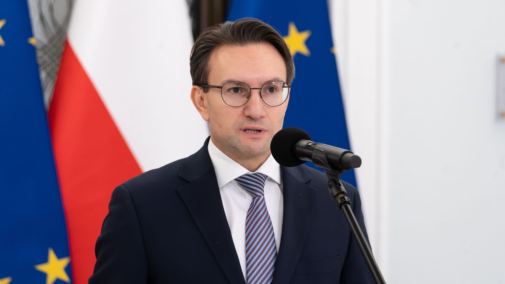 Mężczyzna w garniturze i okularach przemawia do mikrofonu na tle flag Polski i Unii Europejskiej, w kontekście planu tanich mieszkań na wynajem o wartości 45 mld zł do 2030 r.