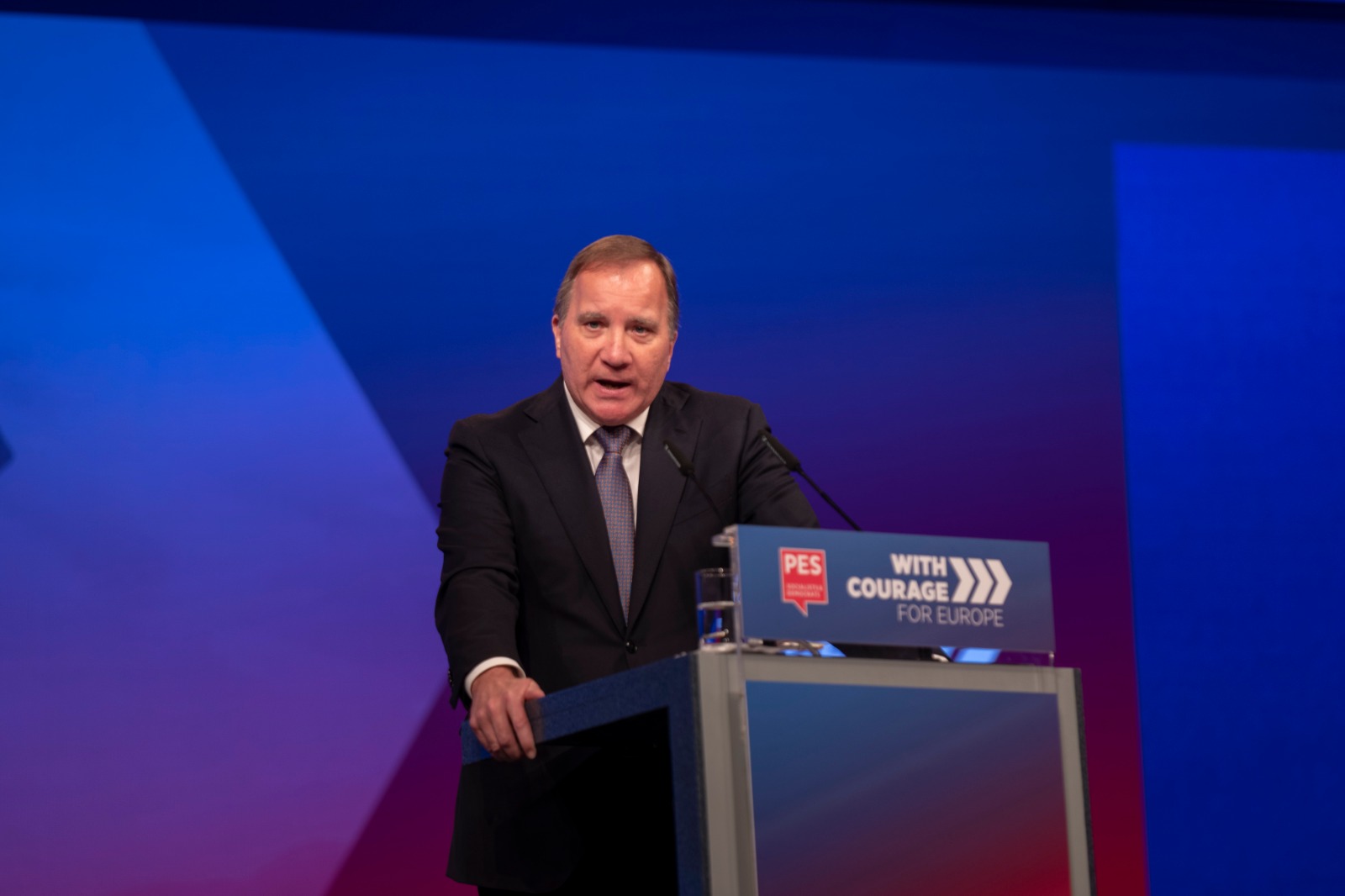Na zdjęciu mężczyzna w garniturze przemawia z mównicy podczas wydarzenia politycznego, z tłem w odcieniach niebieskiego i czerwonego.