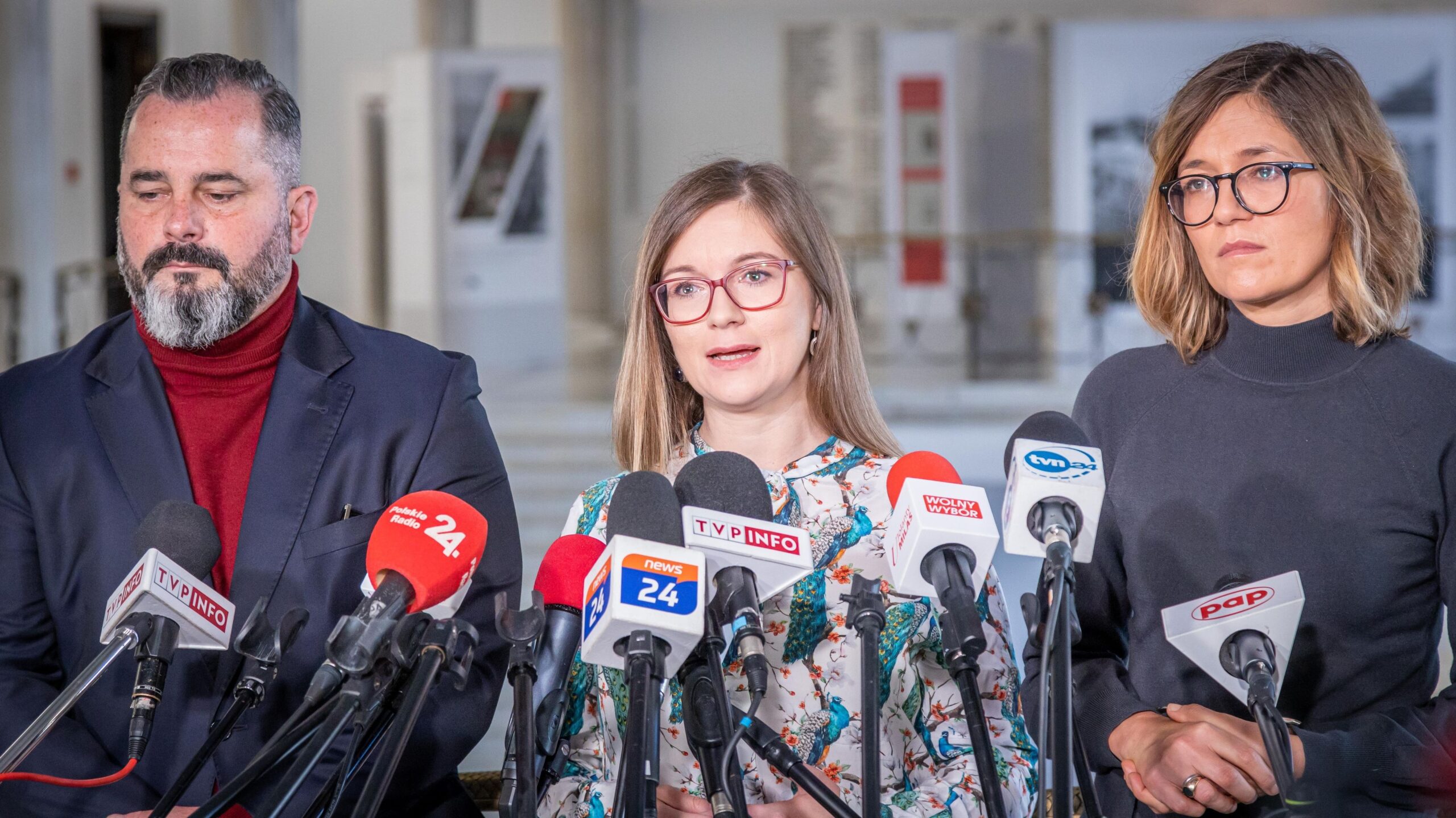 Na zdjęciu widoczna jest trójka osób podczas konferencji prasowej, z mikrofonami i kamerami w tle.