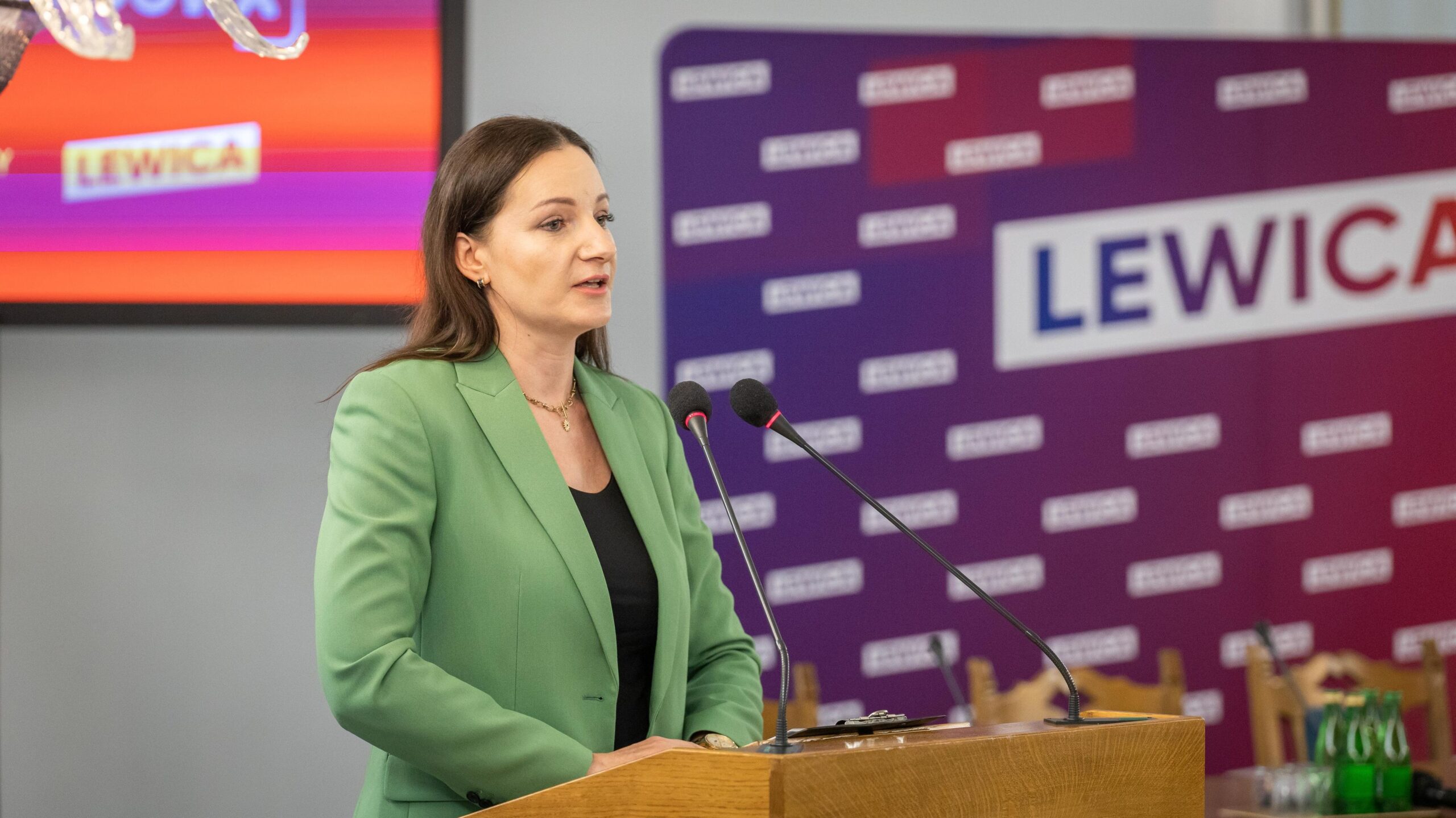 Kobieta w zielonym żakiecie przemawia z mównicy podczas konferencji Lewicy, na tle banerów z napisem LEWICA.