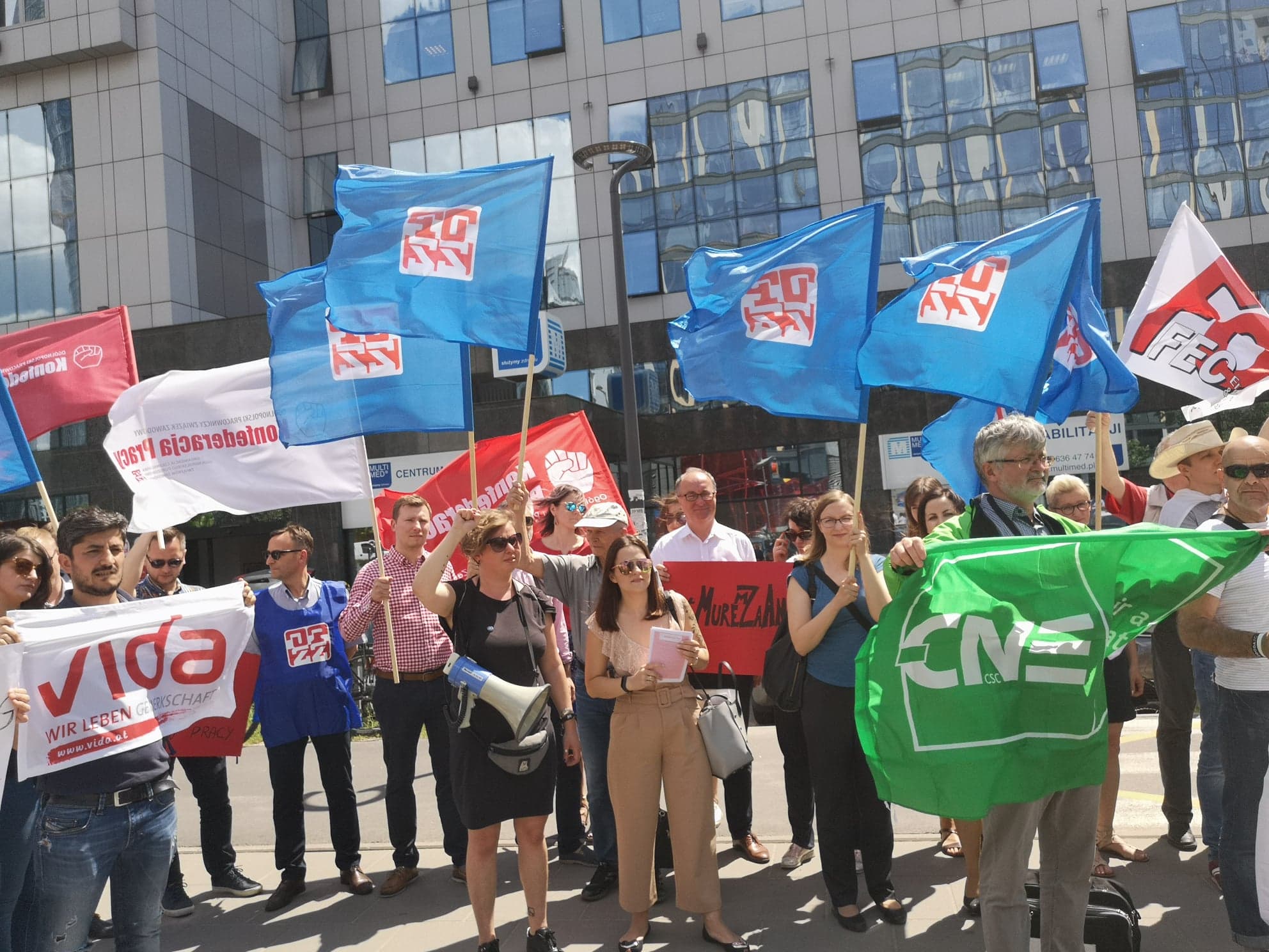 Grupa demonstrantów z flagami i banerami stoi przed nowoczesnym budynkiem, domagając się respektowania konstytucyjnego prawa do zrzeszania się i zakładania związków zawodowych.
