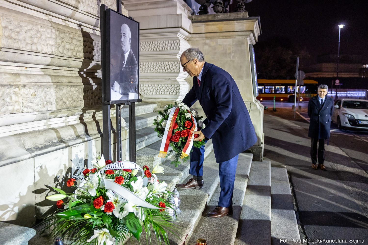 Mężczyzna w ciemnym płaszczu składa wieniec przed portretem prezydenta Narutowicza ustawionym na stojaku przy schodach budynku, w otoczeniu kwiatów, podczas 103. rocznicy zabójstwa.