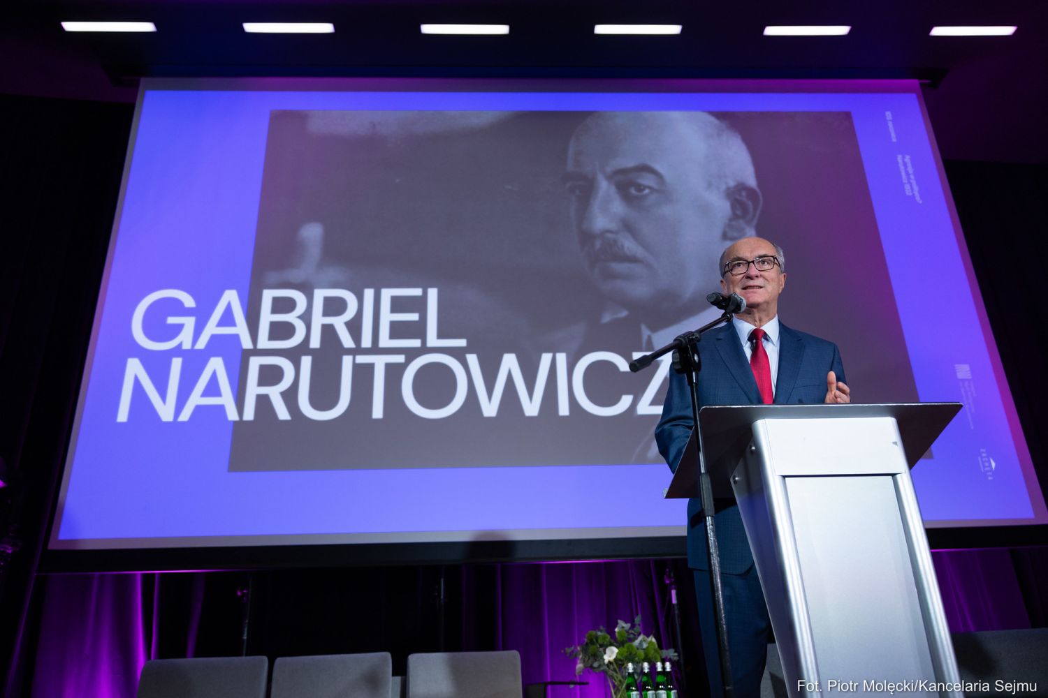 Mężczyzna w garniturze przemawia z mównicy na scenie, a w tle wyświetlany jest duży portret Gabriela Narutowicza i napis upamiętniający 103. rocznicę zabójstwa prezydenta.