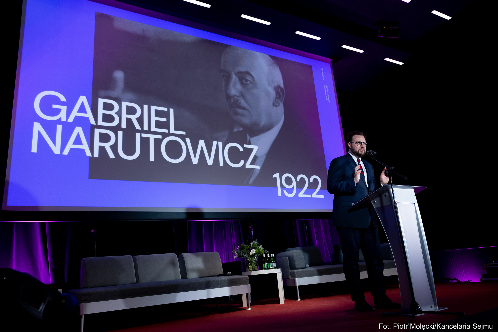 Mężczyzna w garniturze przemawia z podium na scenie podczas konferencji upamiętniającej zabójstwo prezydenta; w tle wyświetlany jest duży ekran z czarno‑białym portretem i napisem odnoszącym się do rocznicy.