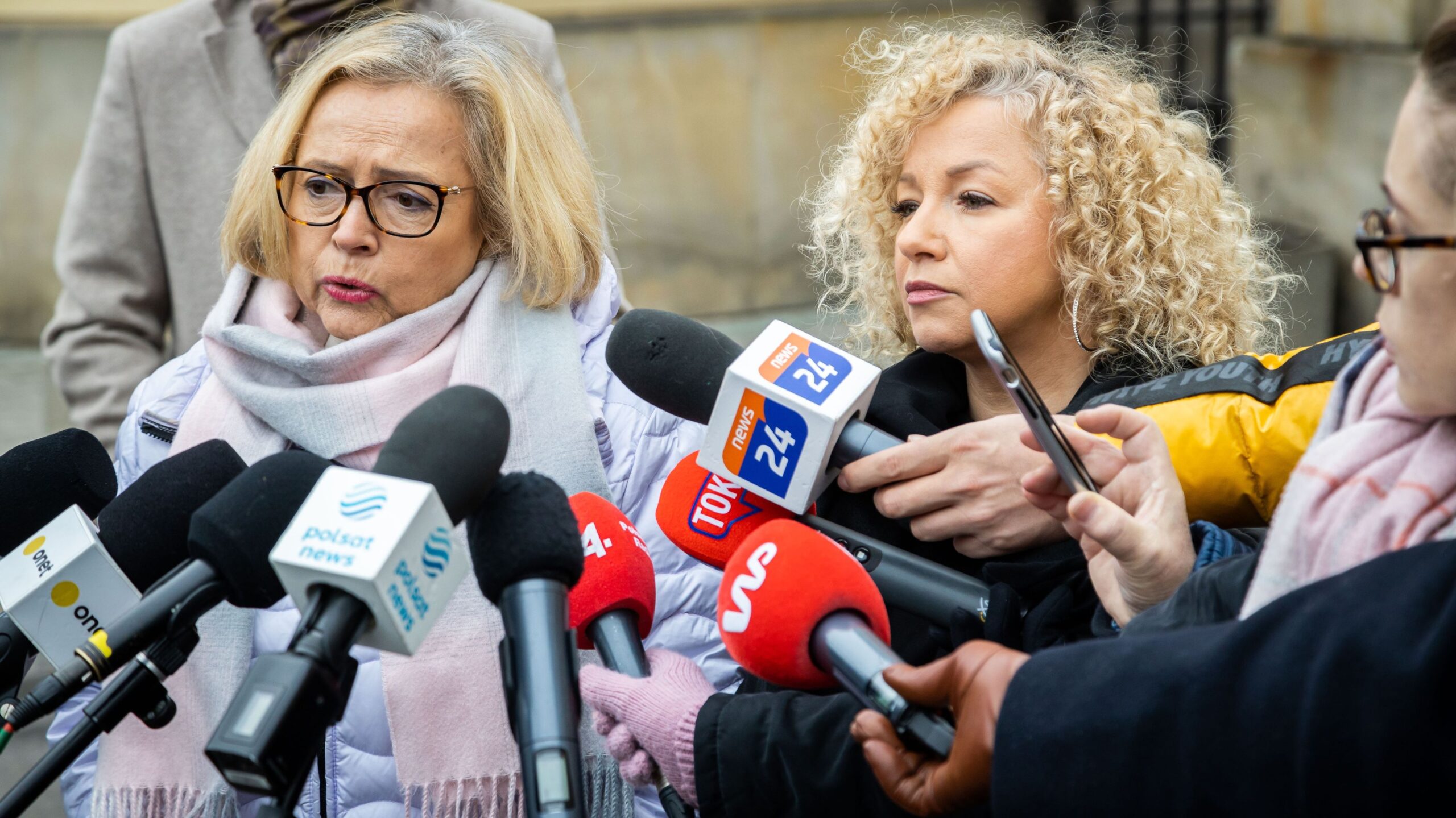 Dwie kobiety udzielają wypowiedzi mediom podczas konferencji prasowej na zewnątrz, otoczone licznymi mikrofonami.