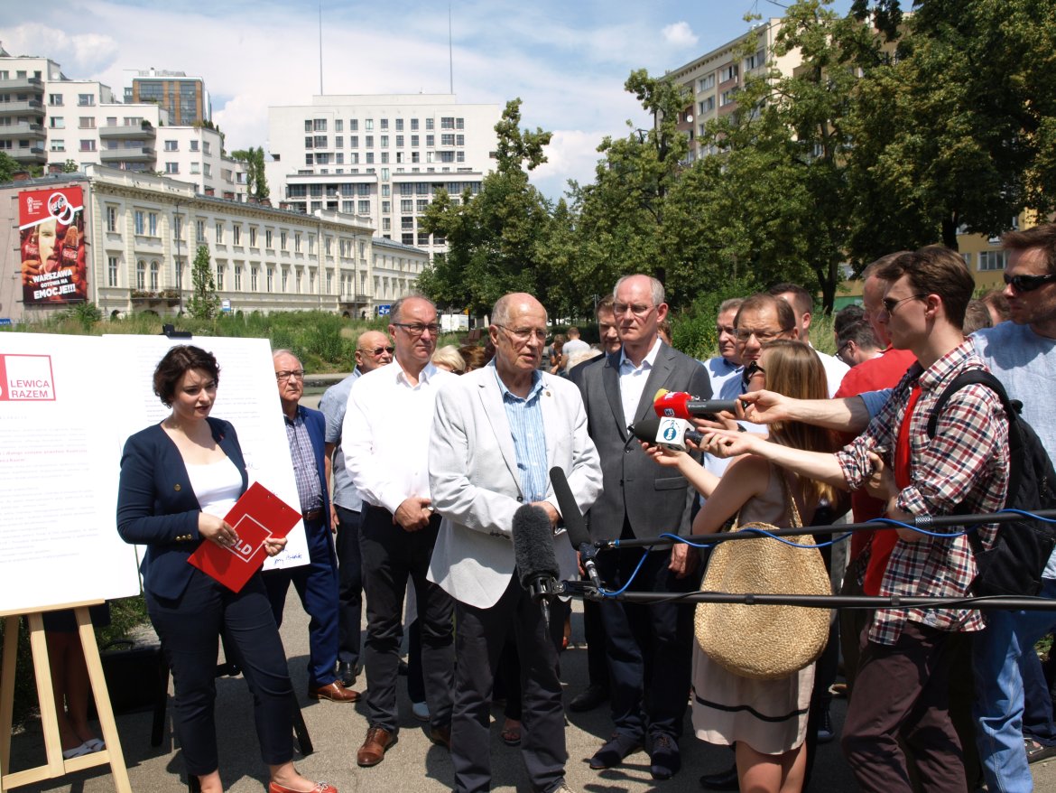 Grupa polityków Lewicy Razem udziela konferencji prasowej na zewnątrz, stojąc obok planszy z logiem Lewicy Razem.