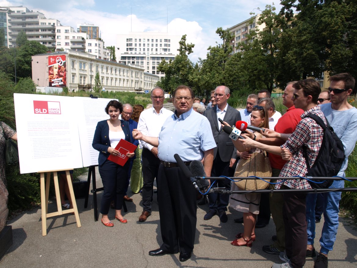 Grupa działaczy SLD – Lewica Razem podczas konferencji prasowej na zewnątrz, z mężczyzną w jasnoniebieskiej koszuli przemawiającym i tablicą informacyjną z logo partii.
