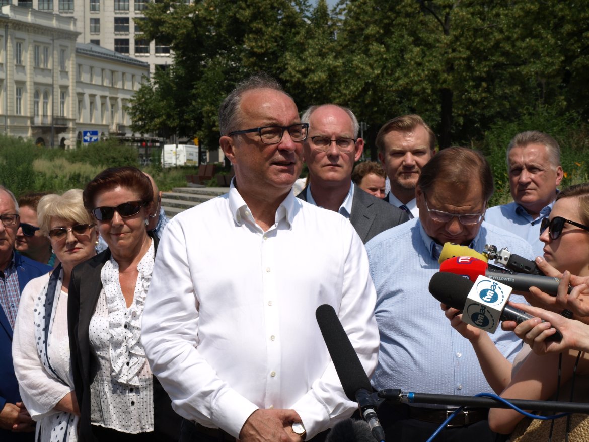Polityk w białej koszuli przemawia do mikrofonów podczas konferencji prasowej na zewnątrz, otoczony przez grupę współpracowników w kontekście koalicji SLD – Lewica Razem.