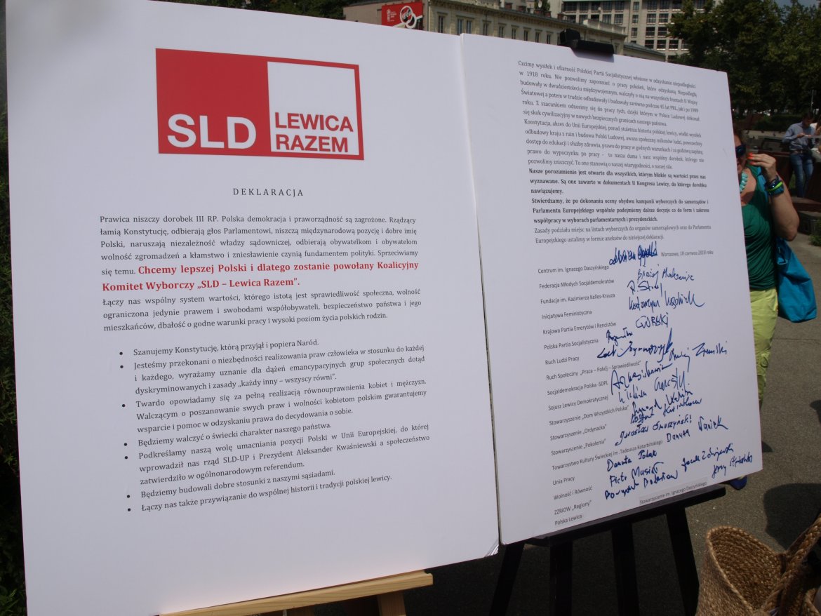 Duży, otwarty plakat na sztaludze z logiem SLD Lewica Razem i deklaracją koalicji, z licznymi podpisami na prawej stronie, ustawiony na zewnątrz.
