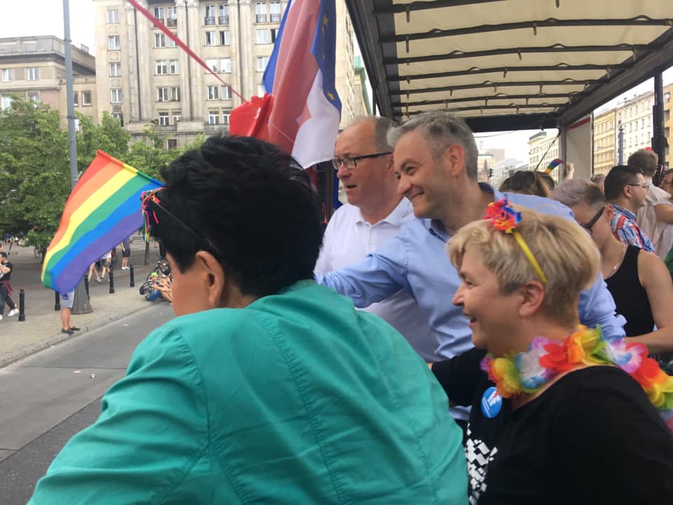 Uczestnicy Parady Równości 2019 stoją na platformie pojazdu na ulicy, trzymają tęczową flagę i noszą kolorowe kwiatowe dodatki.