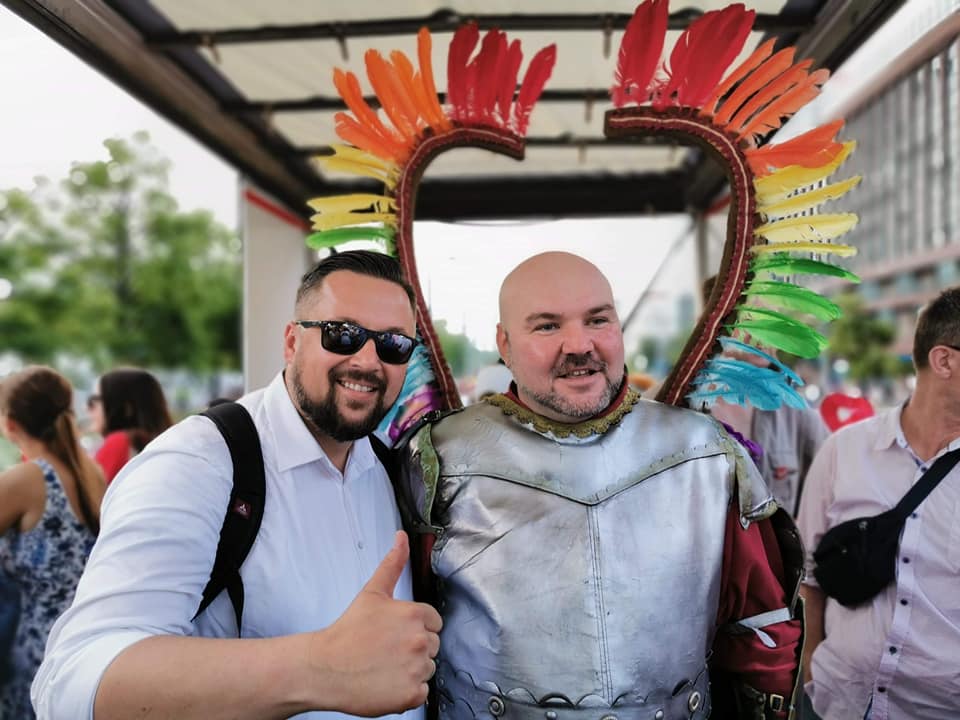Na zdjęciu z Parady Równości 2019 dwóch mężczyzn: jeden w białej koszuli i okularach uśmiecha się i daje kciuk w górę, drugi stoi w błyszczącej srebrnej zbroi pod tęczowym, sercokształtnym obramowaniem z piór.