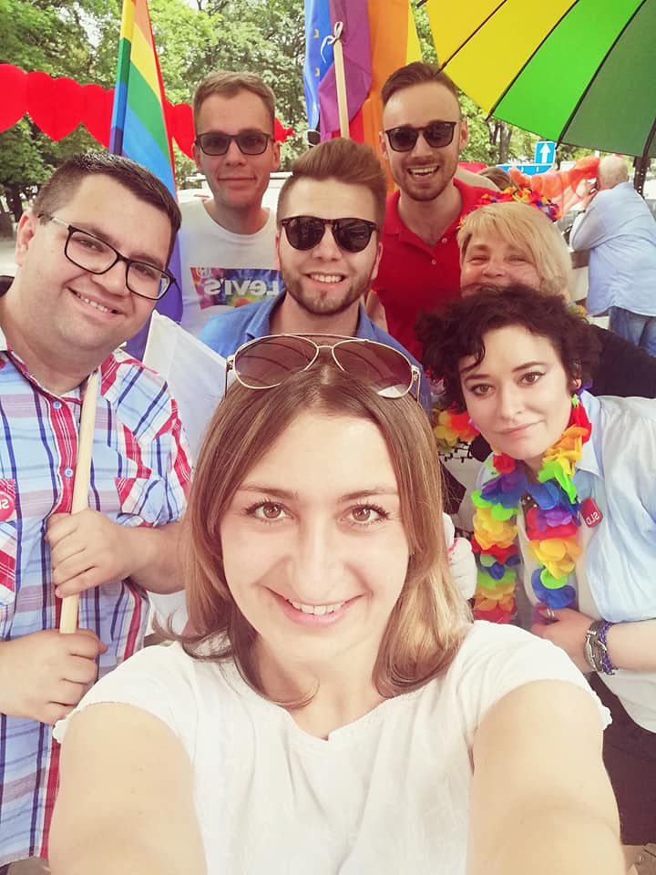 Grupa osób na Paradzie Równości 2019 robi wspólne selfie, w tle widoczne tęczowe flagi i kolorowe parasole.