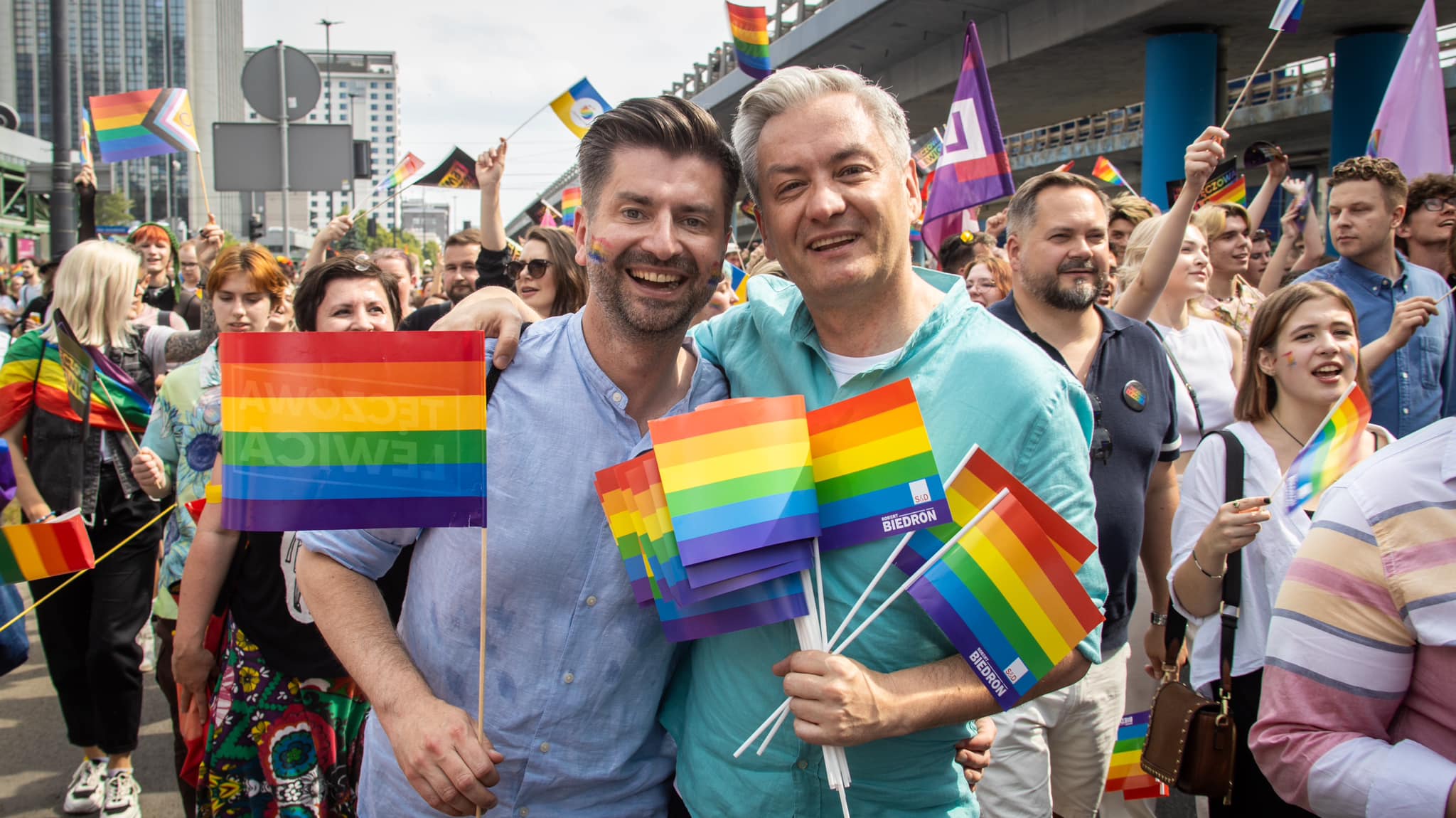 Dwóch mężczyzn stojących ramę w ramię uśmiecha się do kamery podczas Parady Równości 2023, trzymając kolorowe flagi tęczowe w tłumie uczestników.