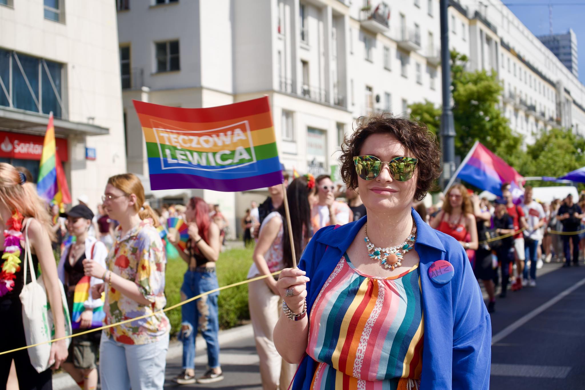 Uśmiechnięta uczestniczka Parady Równości 2024 trzyma flagę w kolorach tęczy z napisem „TĘCZOWA LEWNICA” na ulicy w mieście.