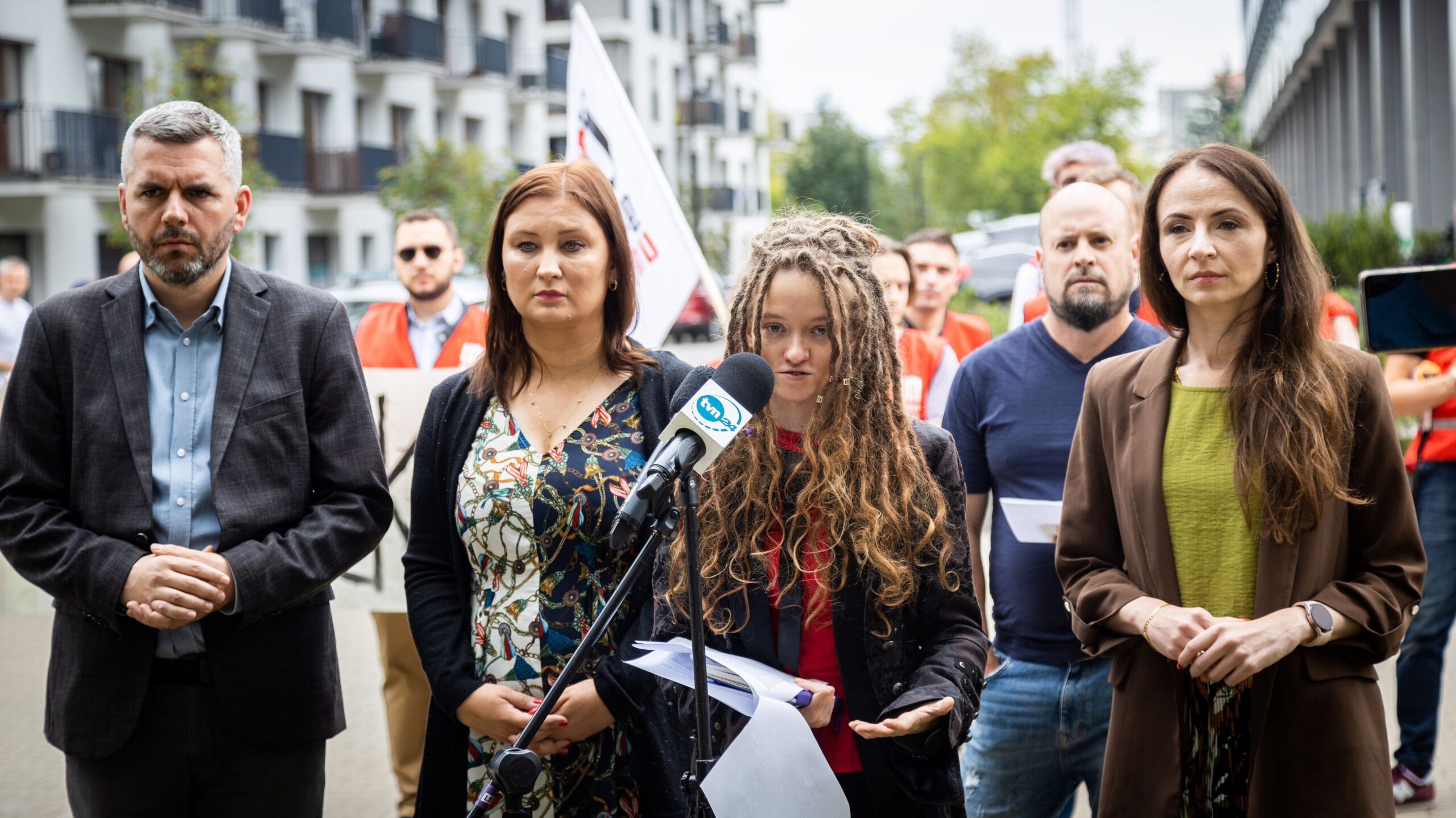 Na zdjęciu przedstawiono grupę osób, z których jedna, w ciemnym płaszczu, mówi do mikrofonu, a pozostałe osoby patrzą w jej stronę, w tle widać budynki i flagi.