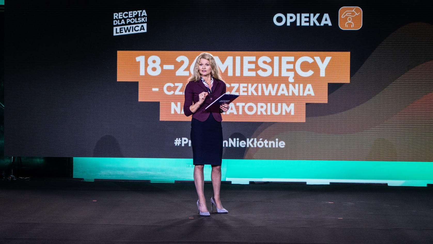 Prezenterka w burgundowej marynarce i czarnej spódnicy stoi na scenie z notatnikiem, a za nią widnieje duży ekran z pomarańczowym napisem i logotypami Recepta dla Polski Lewica oraz Opieka.