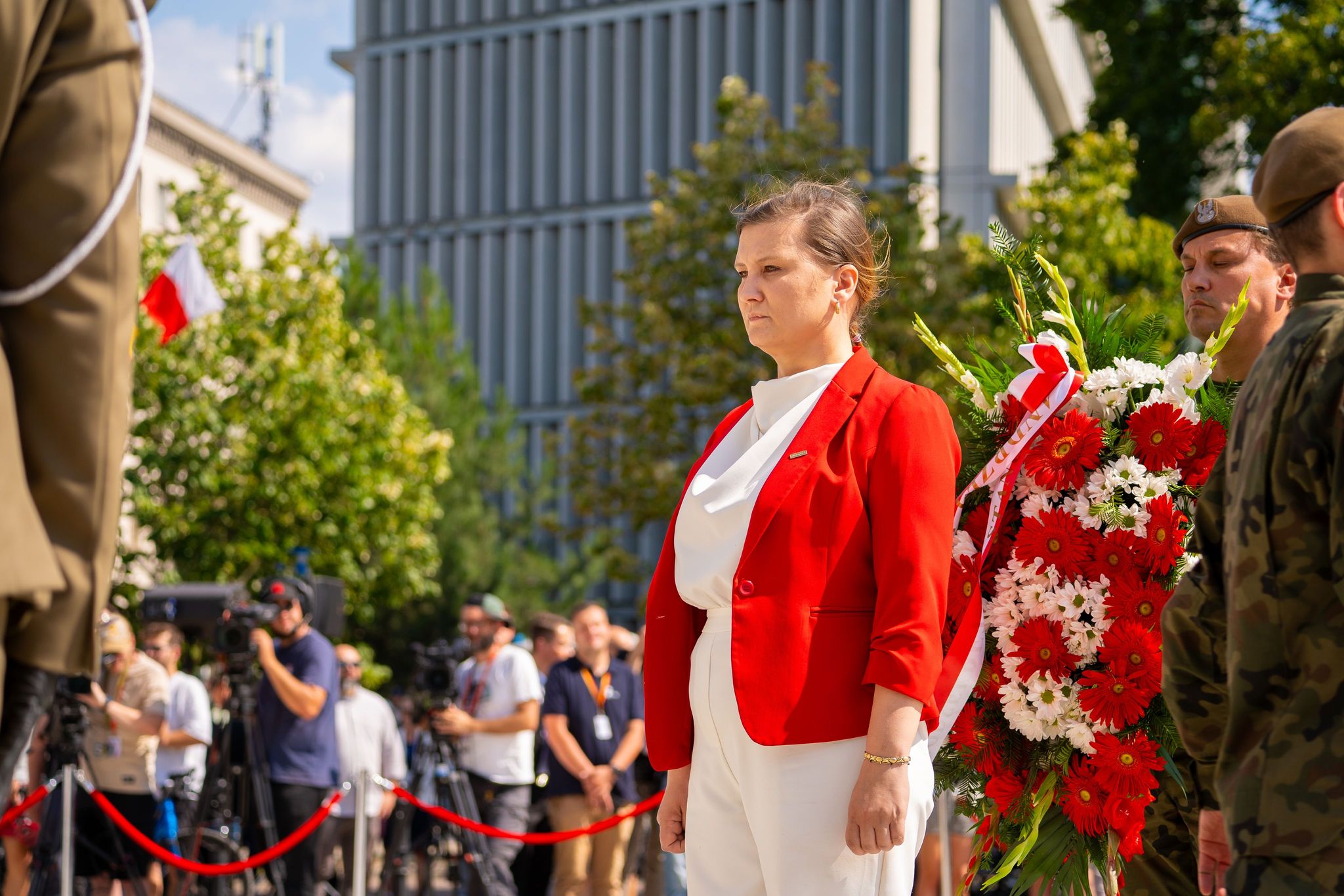 Kobieta w czerwonym żakiecie stoi podczas uroczystości upamiętniającej 80. rocznicę Powstania Warszawskiego, obok dużego wieńca z czerwonych i białych kwiatów, w otoczeniu żołnierzy i fotografów.