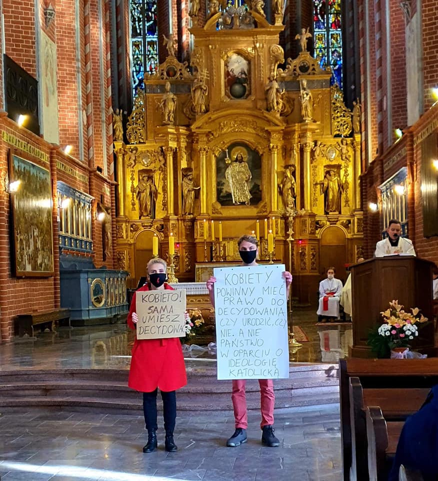 Dwoje demonstrantów z plakatami protestacyjnymi stoi w kościele, podczas gdy duchowny prowadzi nabożeństwo.