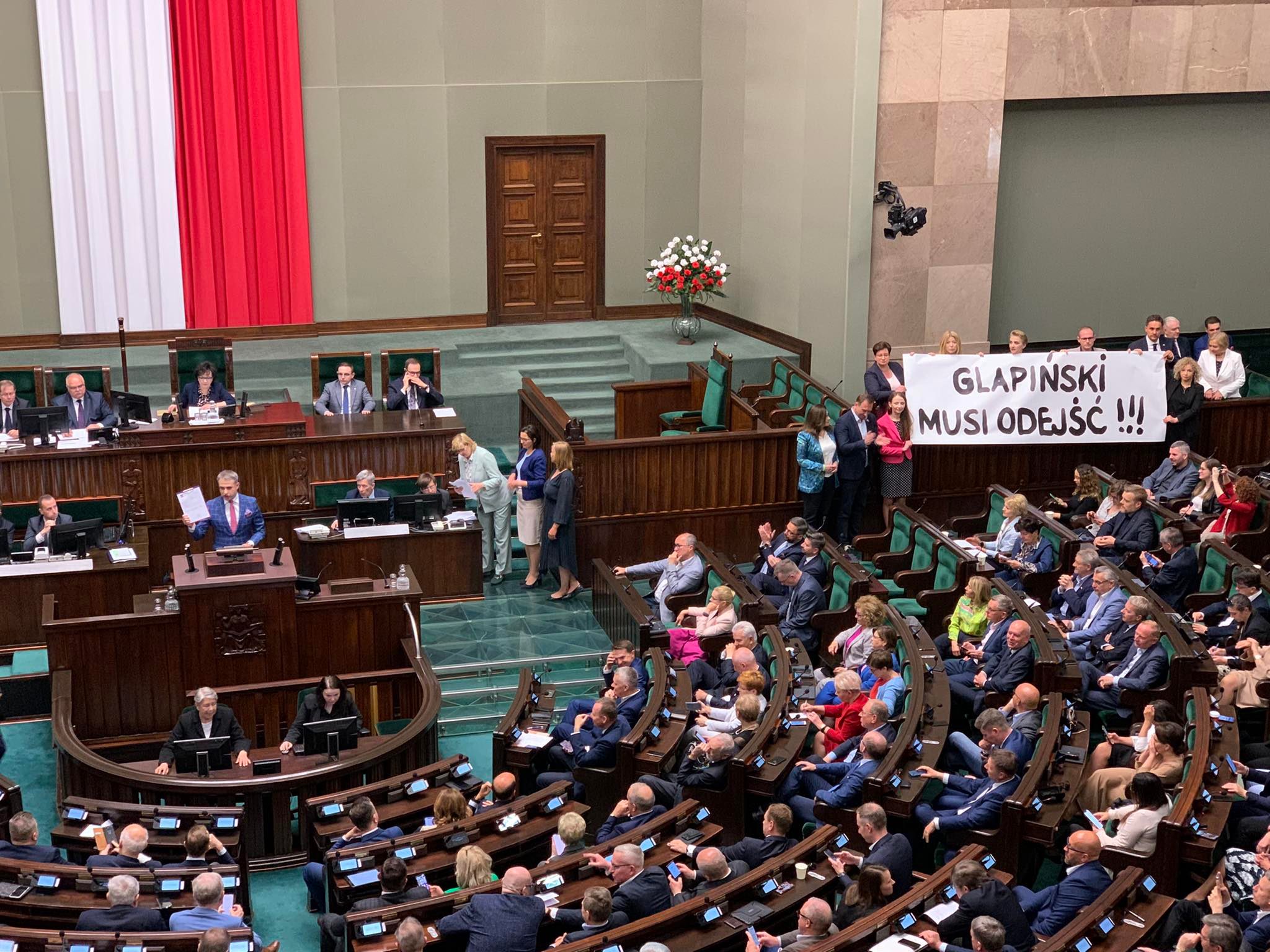 Grupa osób trzyma na galerii sali plenarnej Sejmu duży baner z napisem "GLAPIŃSKI MUSI ODEJŚĆ!!!".