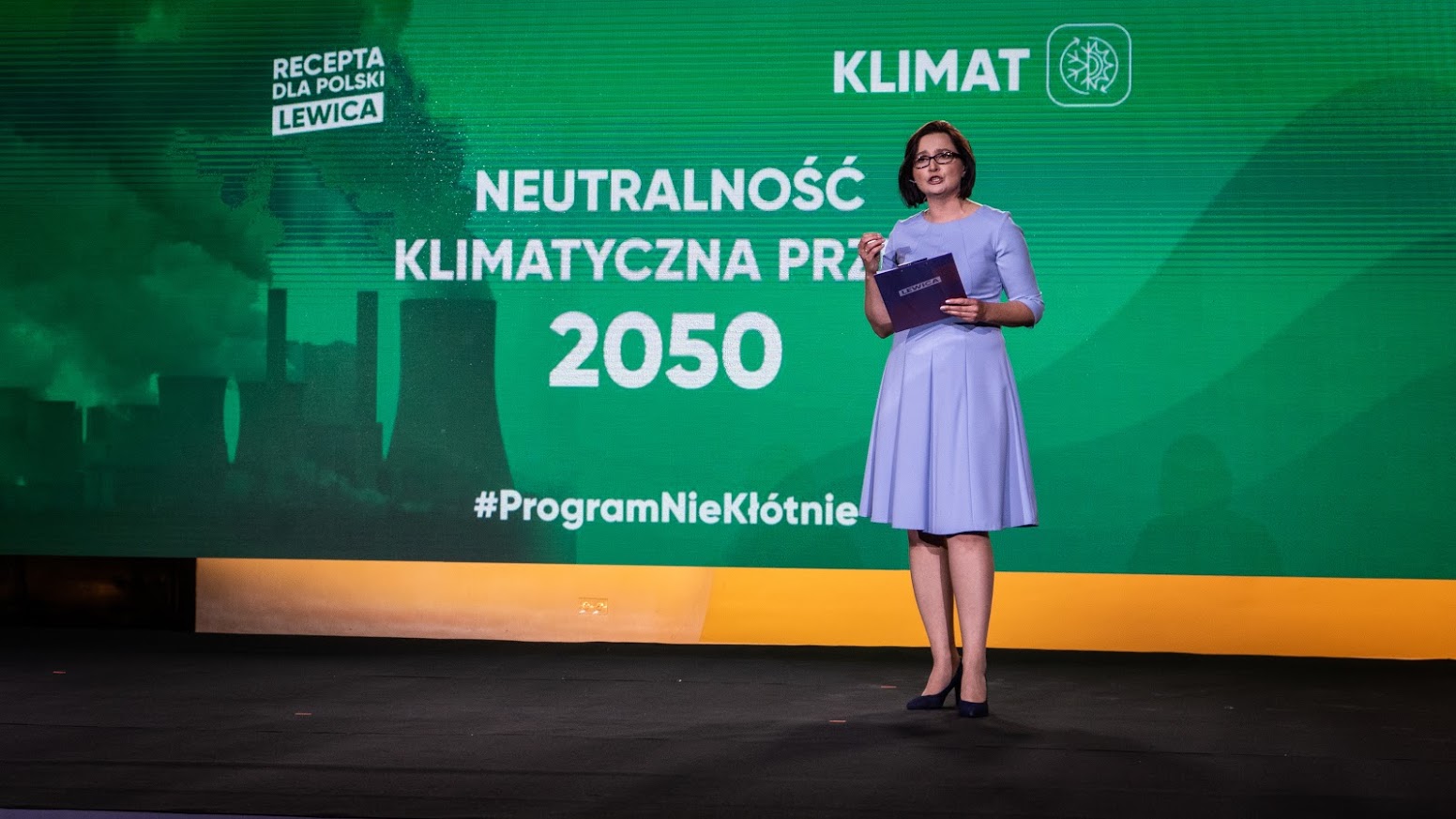 Kobieta stojąca na scenie w jasnoniebieskiej sukience trzyma mikrofon i folder, na tle dużego zielonego baneru z hasłami o neutralności klimatycznej do 2050 roku.