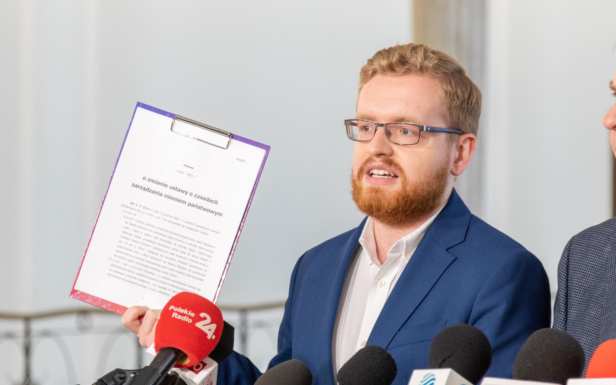 Na zdjęciu widać mężczyznę w garniturze, który podczas konferencji prasowej prezentuje dokument dotyczący zmiany ustawy o zarządzaniu mieniem państwowym.