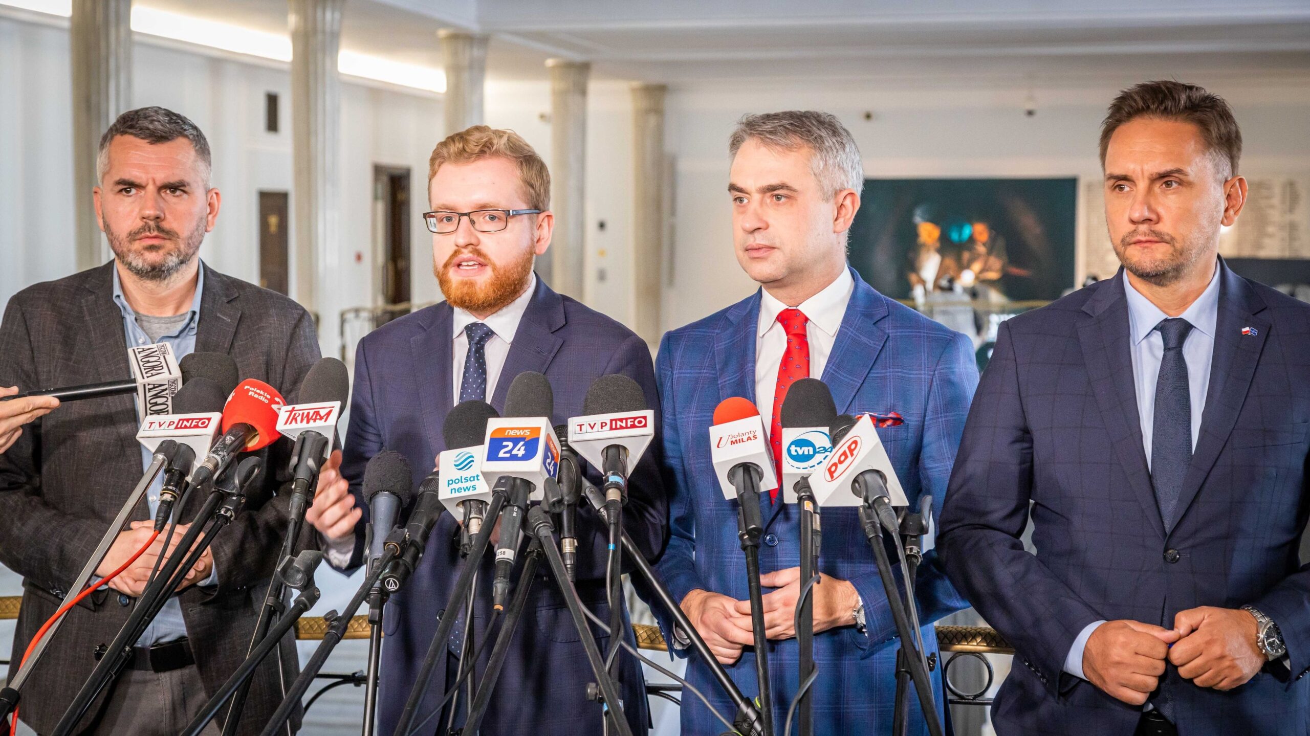 Czterech mężczyzn w formalnych strojach stoi przy mikrofonach podczas konferencji prasowej w jasnym wnętrzu.