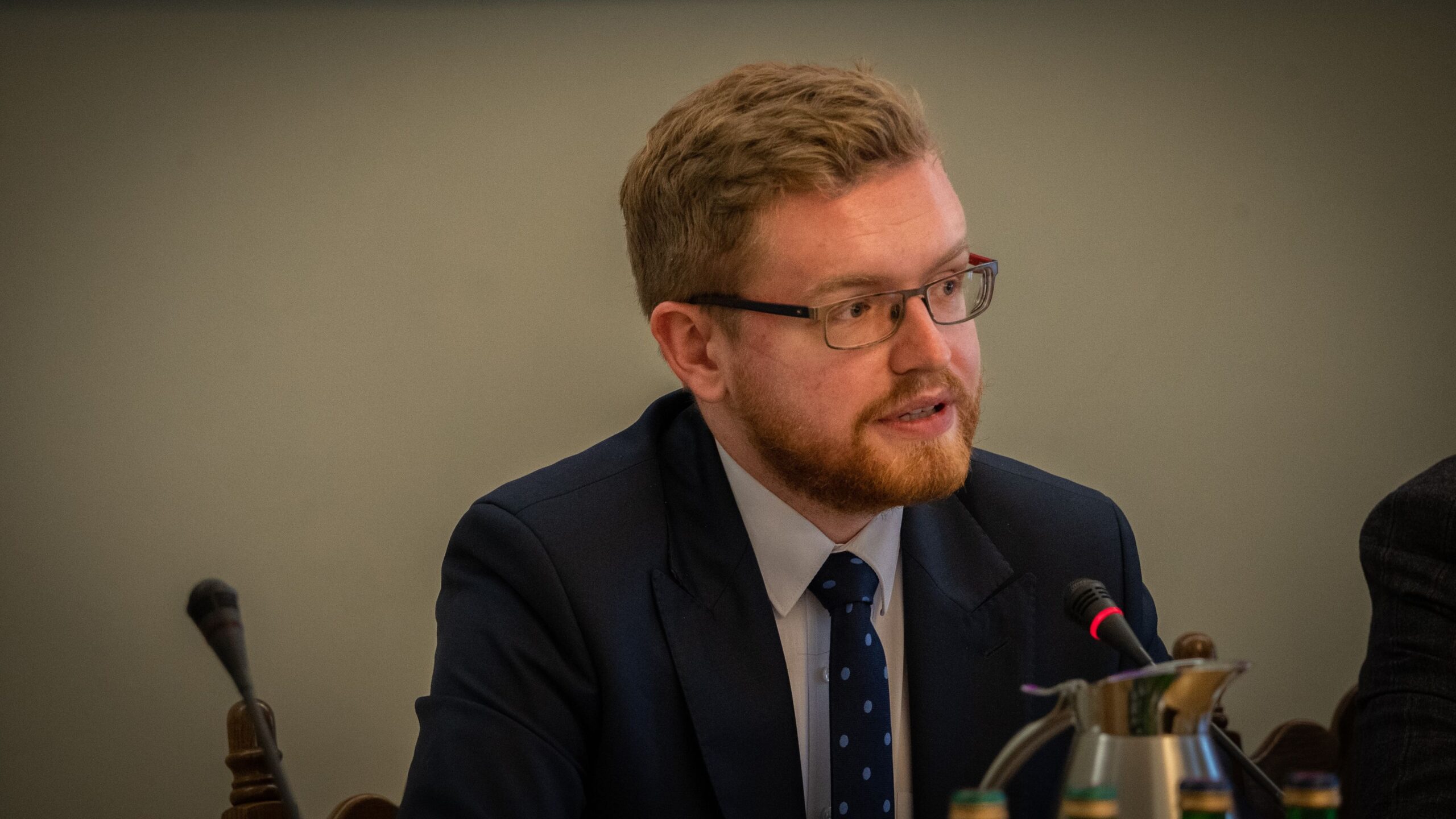 Mężczyzna w garniturze z okularami, z brodą i włosami w odcieniach blond, rozmawia podczas spotkania, siedząc za stołem.