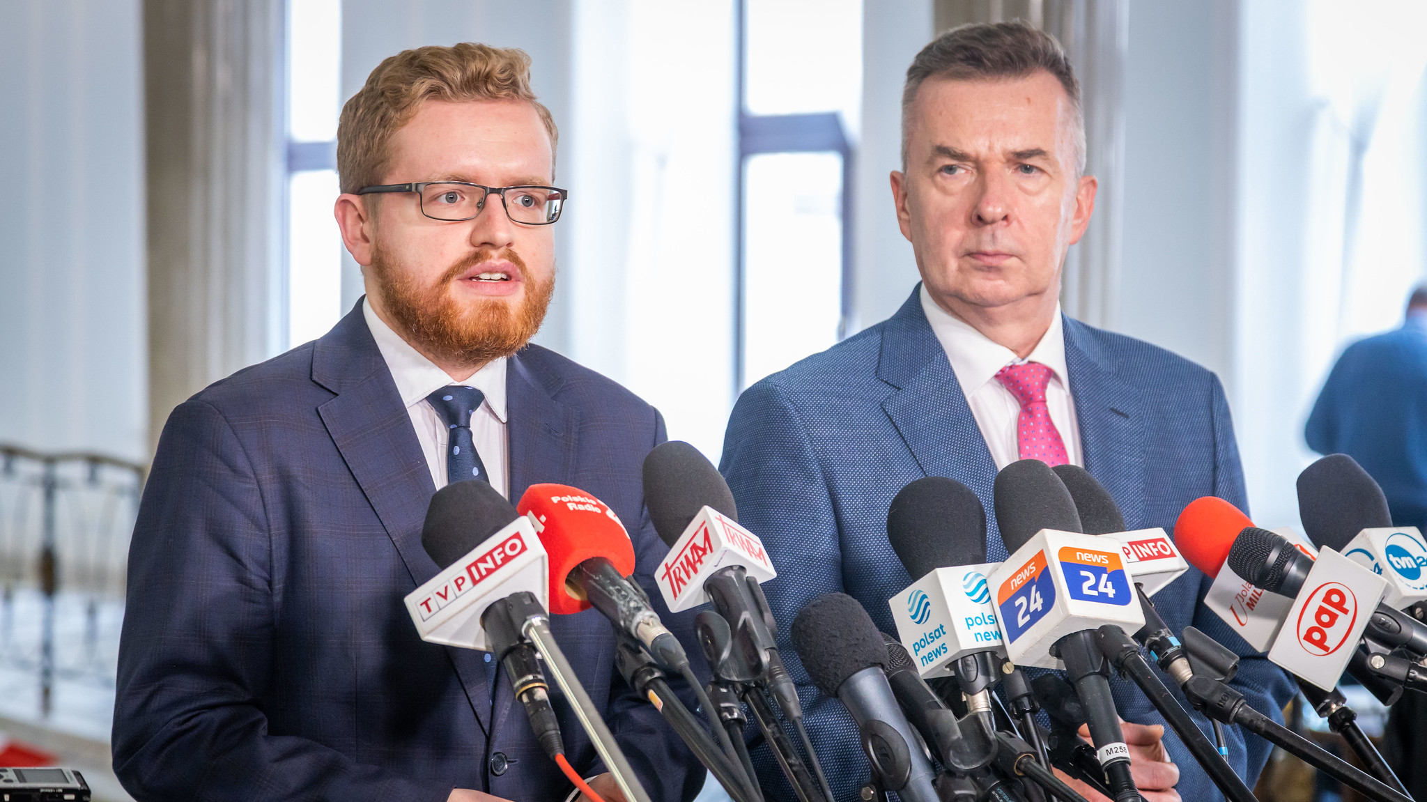 Na zdjęciu dwóch mężczyzn w garniturach stoi za mikrofonami podczas konferencji prasowej w budynku publicznym.