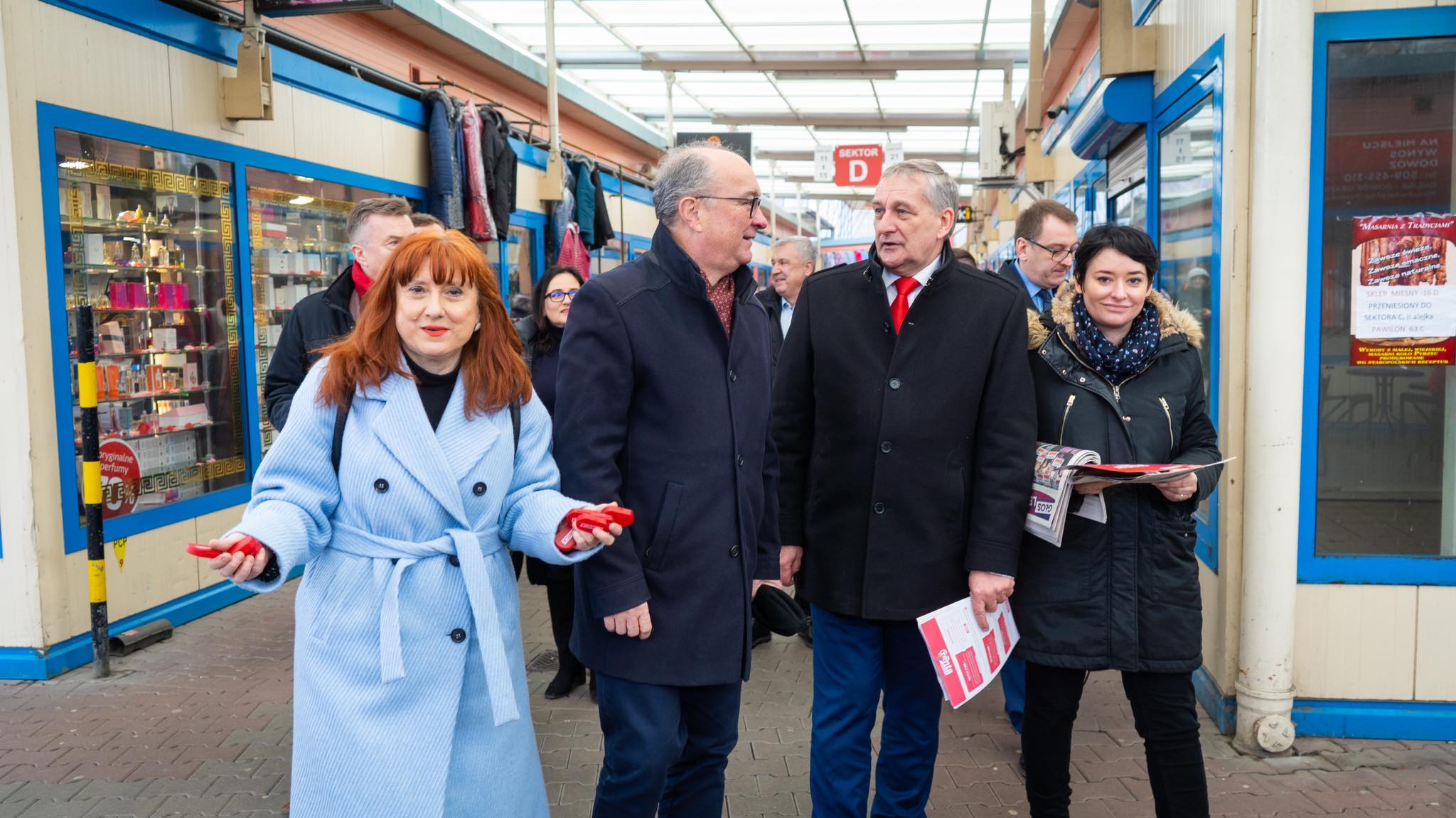 Grupa osób w zimowych płaszczach spaceruje po przejściu targowym w Szczecinie; na pierwszym planie uśmiecha się kobieta w jasnoniebieskim płaszczu, podczas gdy pozostali trzymają ulotki i rozmawiają.