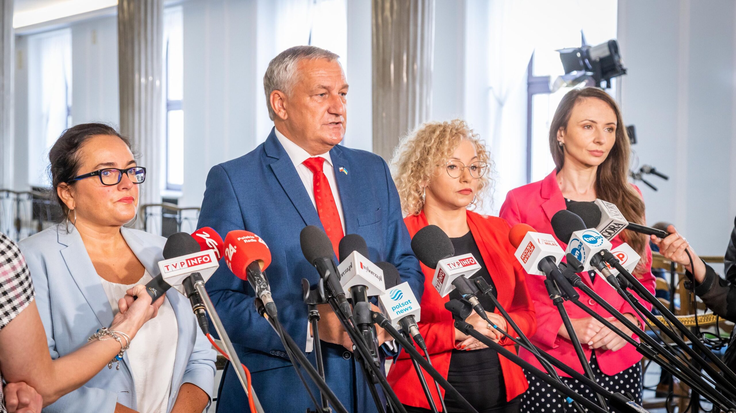Grupa polityków na konferencji prasowej, stojąca za licznymi mikrofonami z logotypami mediów w sali konferencyjnej.