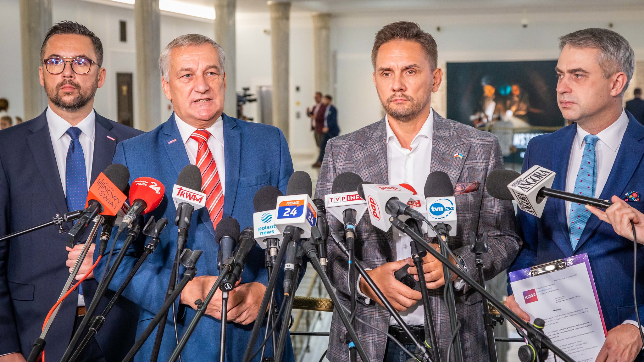 Na zdjęciu widoczna jest grupa czterech mężczyzn podczas konferencji prasowej, otoczonych mikrofonami i kamerami w tle.