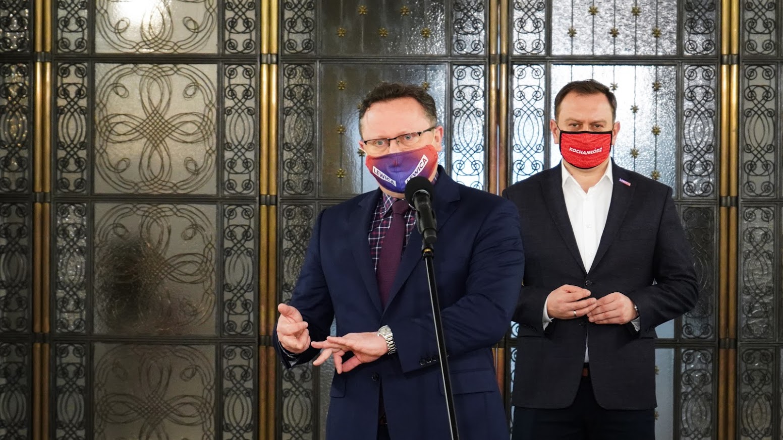 Dwóch mężczyzn w garniturach prowadzi konferencję prasową, jeden mówi do mikrofonu, drugi stoi obok, oboje mają maseczki z politycznymi napisami, a w tle widoczne ozdobne drzwi.