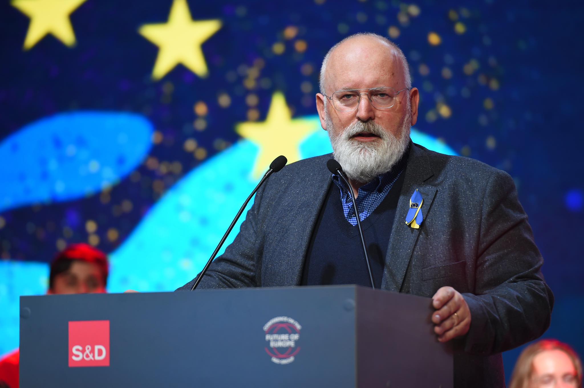 Na zdjęciu widać mężczyznę z brodą, mówiącego z mównicy na tle kolorowego tła, symbolizującego Unię Europejską.