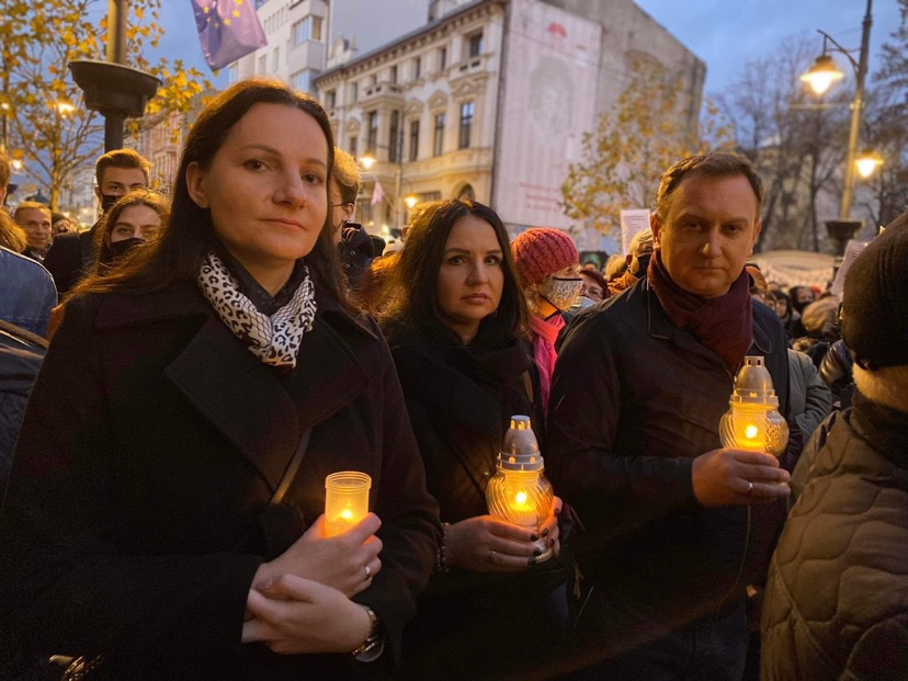 Grupa ludzi trzymających świece i znicze, zgromadzona na miejskim placu w czasie wiecu.