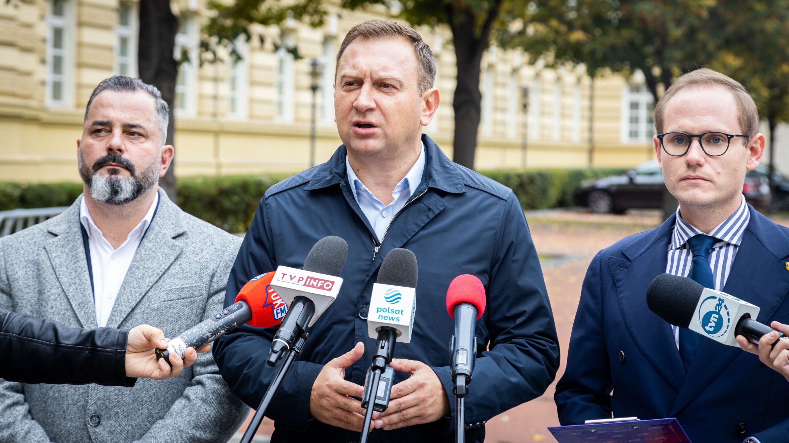 Trzech mężczyzn w garniturach podczas konferencji prasowej na świeżym powietrzu, z mikrofonami i dziennikarzami w tle.