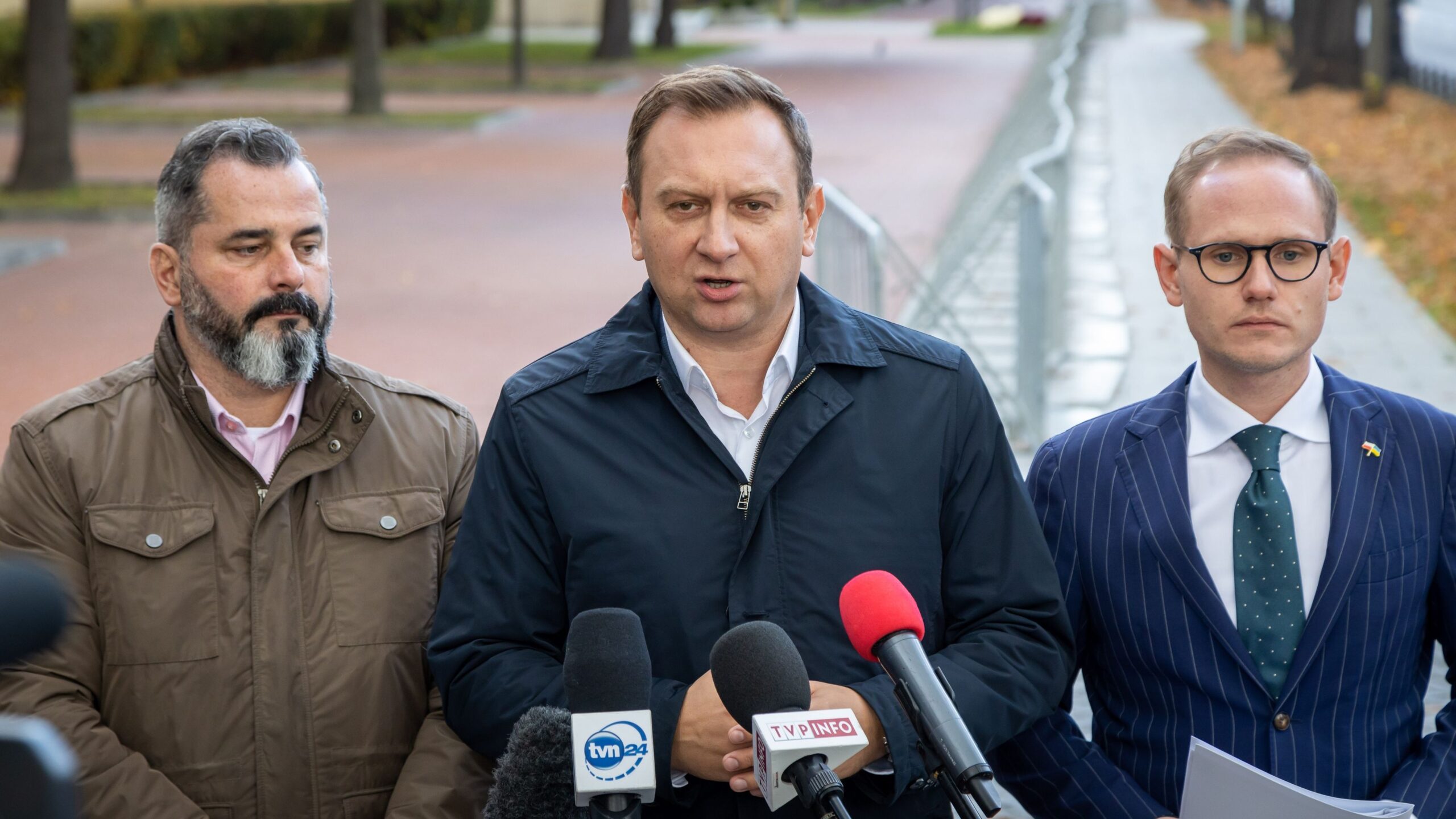 Na zdjęciu trzej mężczyźni stoją przed mikrofonami podczas konferencji prasowej na zewnątrz, w otoczeniu miejskim.