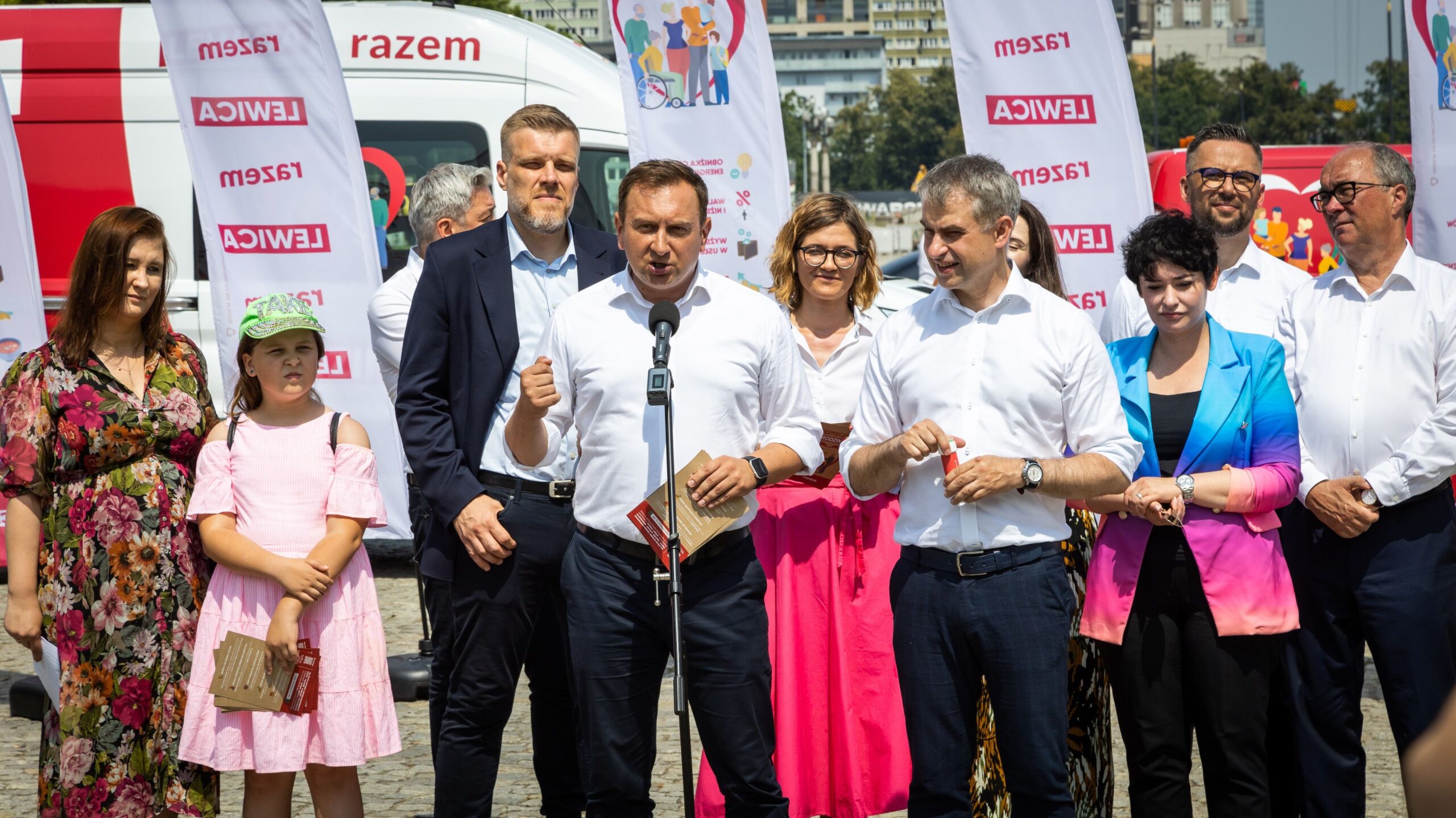 Grupa osób stojących na plenerowej konferencji prasowej podczas wydarzenia „Wakacyjna trasa Lewicy – Bezpieczna Rodzina”, z mężczyzną w białej koszuli mówiącym do mikrofonu, otoczonych współorganizatorami i dziewczynką w różowej sukience, z widocznymi flagami Lewicy w tle.