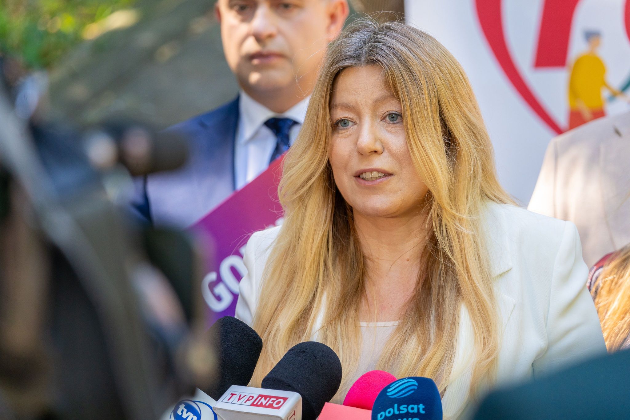 Kobieta z blond włosami w jasnej marynarce przemawia do mikrofonów podczas konferencji prasowej, na tle banneru.