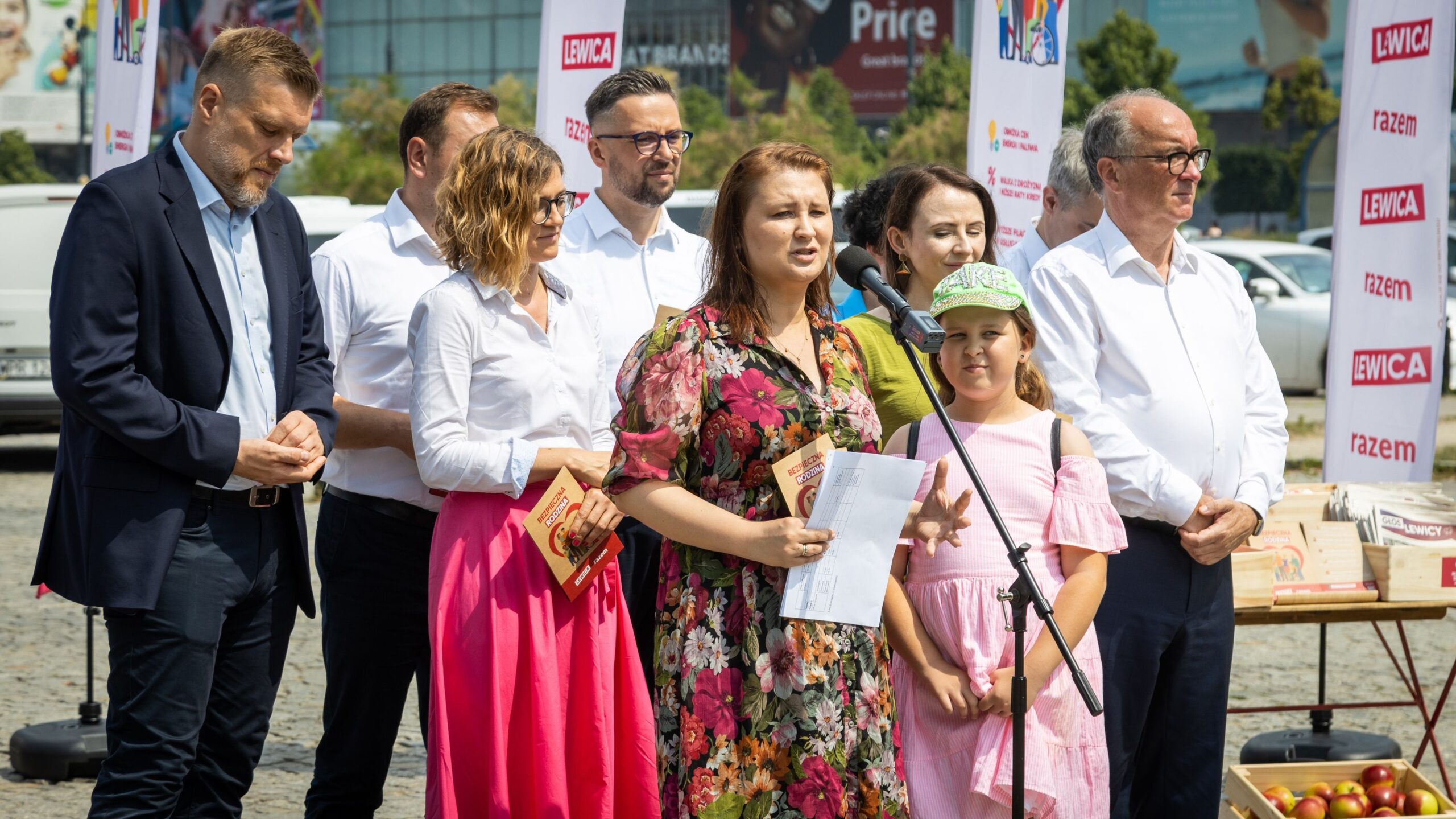 Na zdjęciu przedstawiono grupę osób, w tym kobietę przemawiającą do mikrofonu, z uśmiechniętymi dziećmi i innymi uczestnikami, w otoczeniu banerów politycznych podczas wydarzenia na świeżym powietrzu.