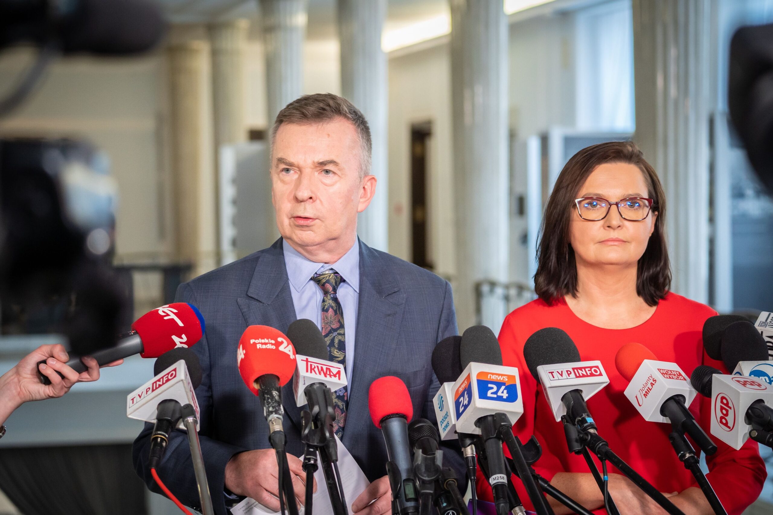 Dwóch polityków stoi za mikrofonami podczas konferencji prasowej w przestronnej sali.
