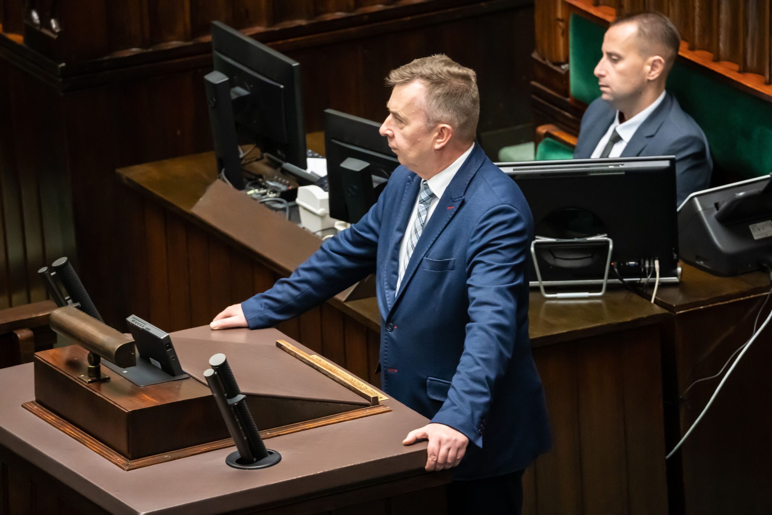 Na zdjęciu widać mężczyznę w garniturze przemawiającego z mównicy w sali obrad, a w tle widać innych uczestników sesji.