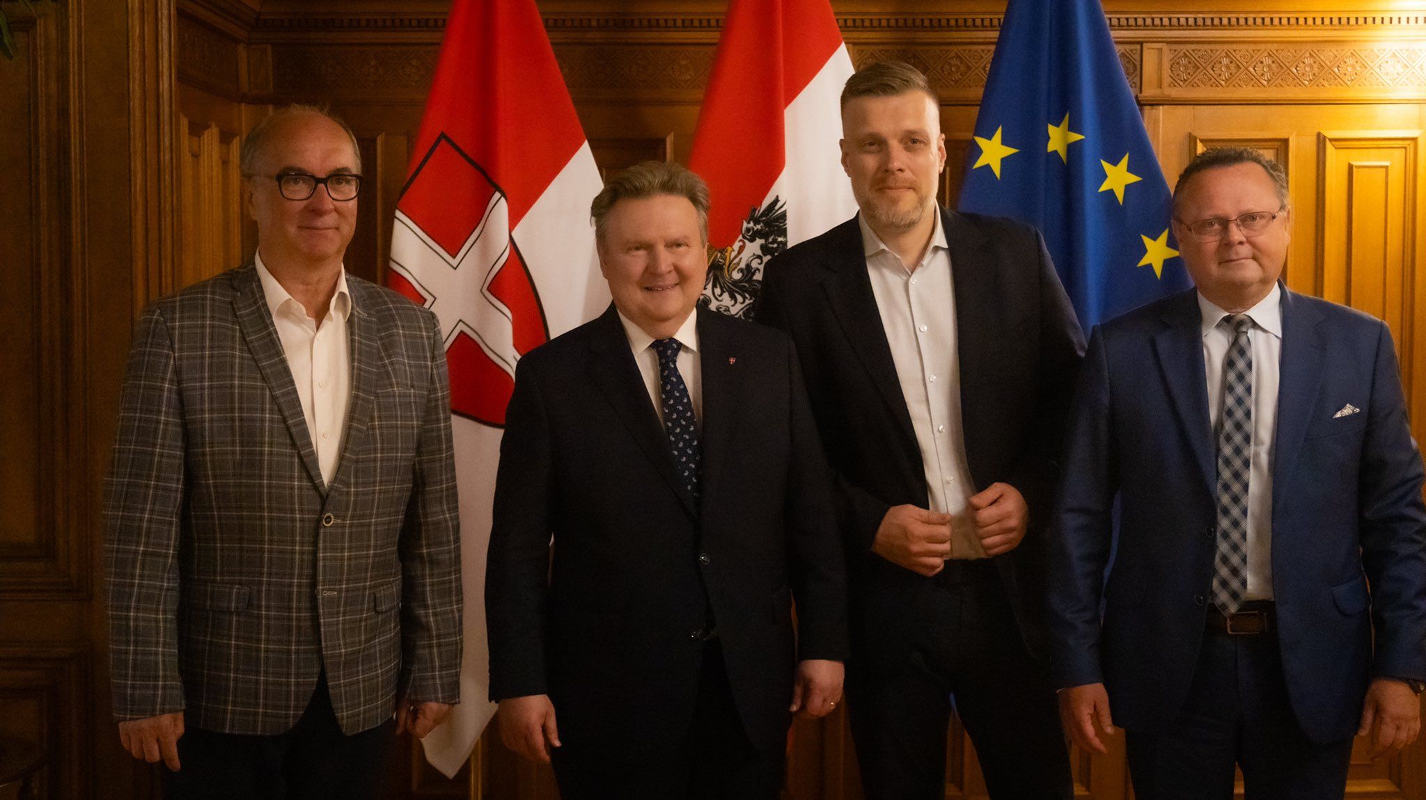 Czterech mężczyzn w garniturach stoi w drewnianej sali z flagami w tle, sugerując oficjalne spotkanie.