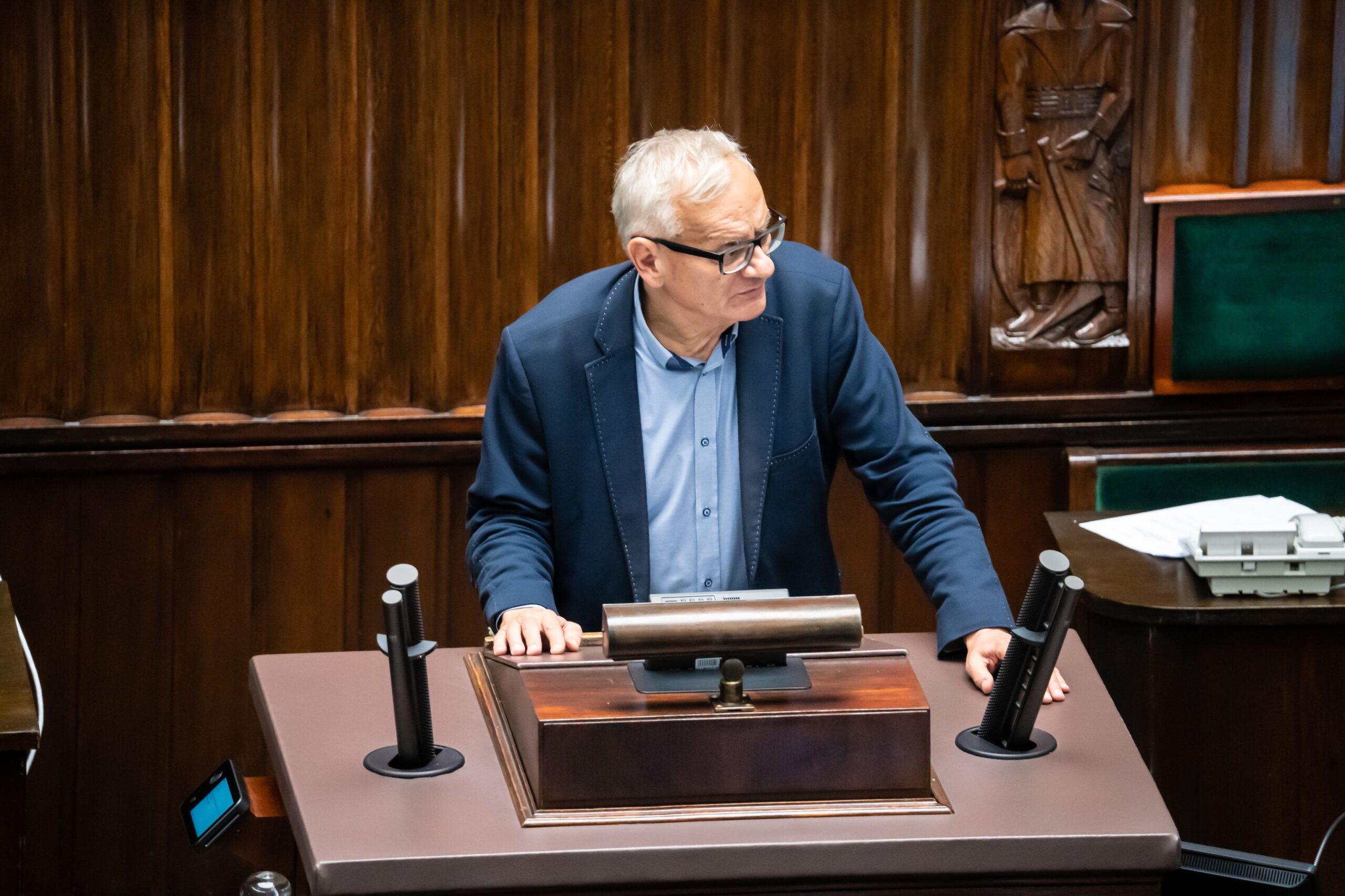 Na zdjęciu widać mężczyznę w okularach, stojącego przy mównicy w parlamencie, który wygląda na zaangażowanego w dyskusję.