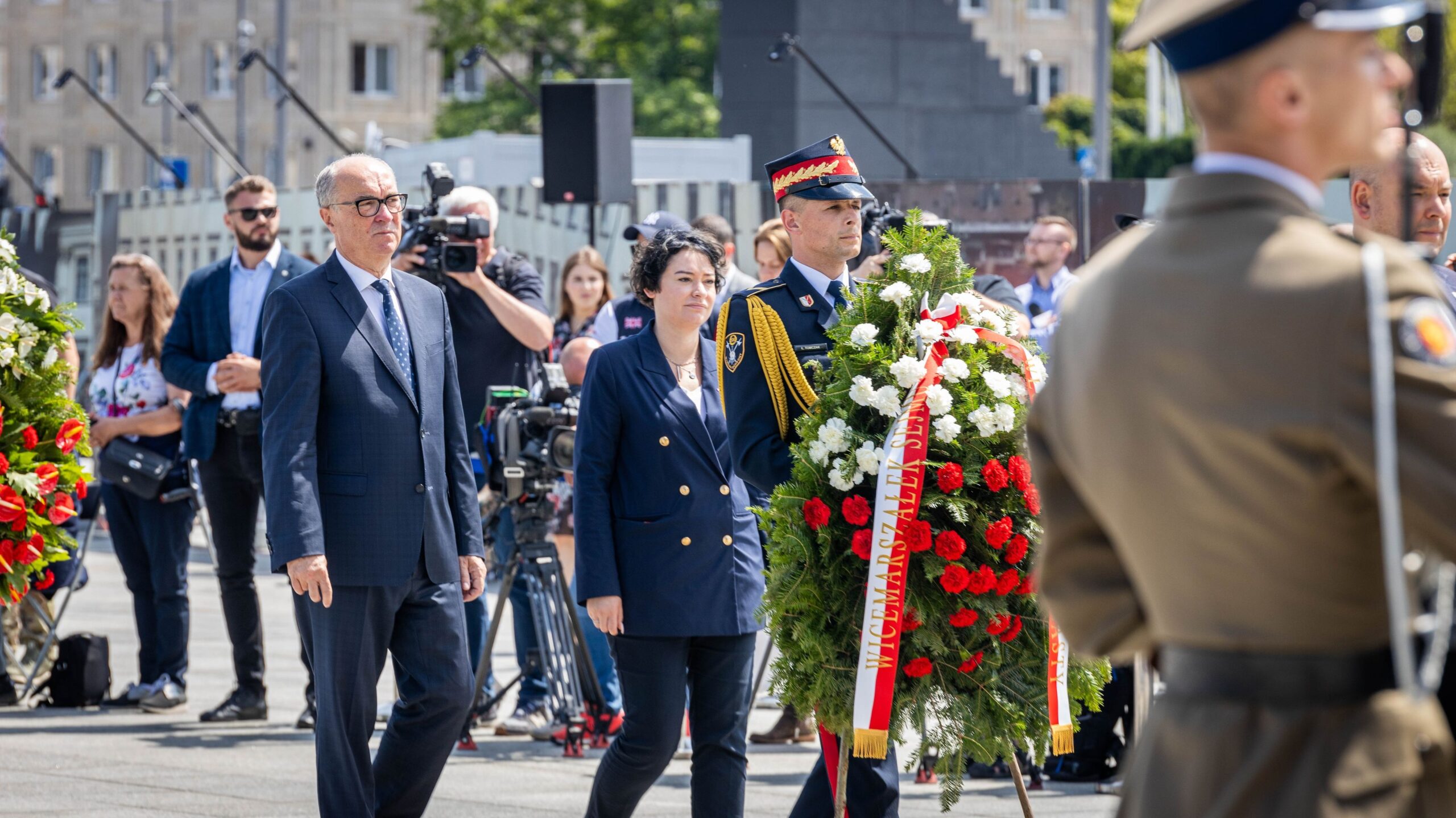 Uczestnicy uroczystości upamiętniającej grob Nieznanego Żołnierza stoją na placu, a w centrum kadru widoczny jest starszy mężczyzna w garniturze, kobieta w granatowym garniturze i żołnierz w mundurze niosący duży wieniec z białych i czerwonych kwiatów, otoczeni przez reporterów i innych uczestników.