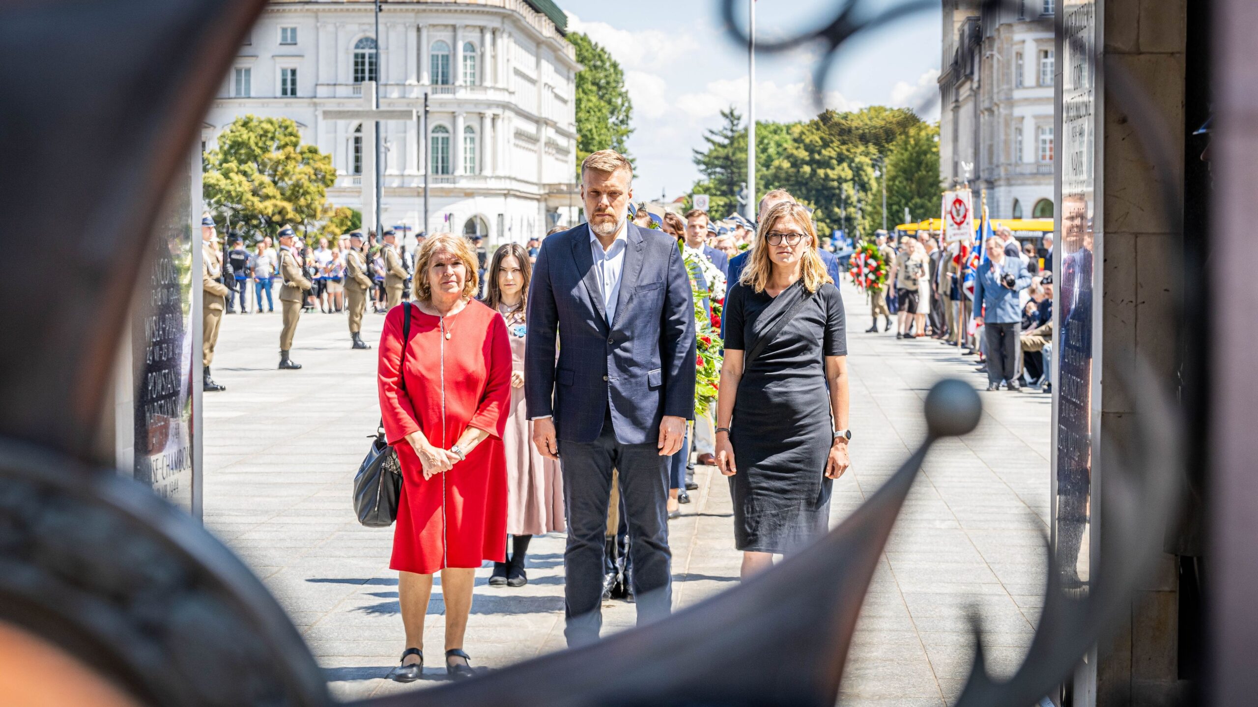 Grupa uczestników ceremonii stoi na placu przed pomnikiem grobu Nieznanego Żołnierza, w tle widoczni żołnierze i zgromadzeni ludzie.