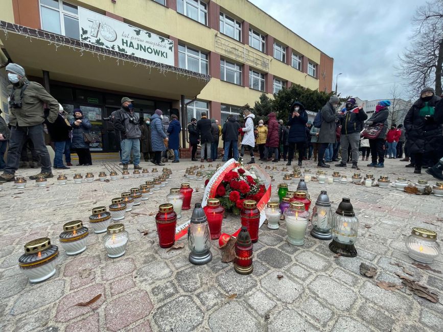 Obraz przedstawia grupę ludzi zgromadzonych przed budynkiem w Hajnowce, wokół symbolicznych zniczy i wieńca z czerwonymi różami, w atmosferze zadumy.