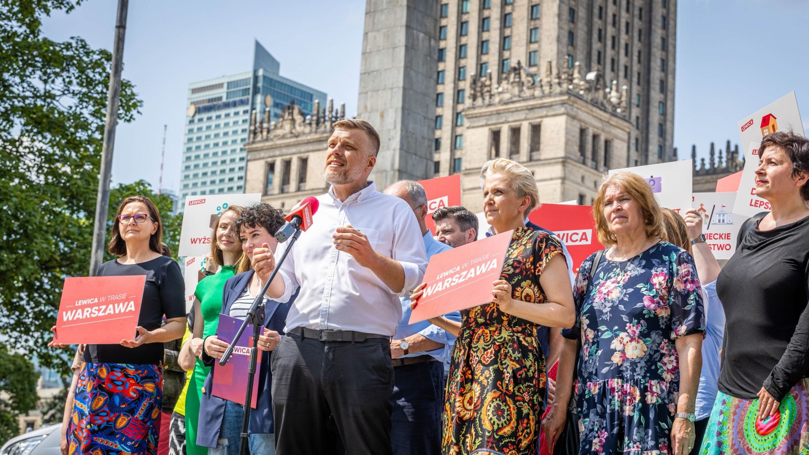 Grupa działaczy Lewicy podczas plenerowego wystąpienia w Warszawie, mężczyzna w białej koszuli przemawia do mikrofonu, otoczony czerwonymi tablicami z napisem Warszawa.