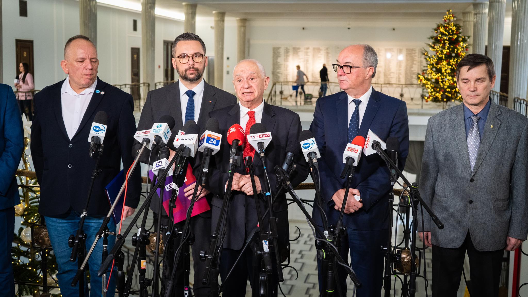 Na zdjęciu widać grupę mężczyzn stojących przy mikrofonach na konferencji prasowej, w tle choinka bożonarodzeniowa.